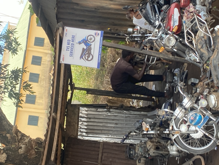 This image captures an outdoor motorcycle repair workshop in Afikpo, Nigeria. The scene is dominated by a makeshift structure with a corrugated metal roof and walls, supported by wooden poles. A prominent white rectangular sign, suspended from a wooden beam, advertises "TVS NEO SERVICED HERE" with an image of a blue motorcycle and states "COMPANY TRAINED TECHNICIANS".
Two men are actively working on motorcycles. The man in the foreground, dressed in a dark shirt and trousers, is bent over a motorcycle, performing mechanical work on its engine or chassis. To his right and slightly behind, a second man in a white sleeveless top and cap is similarly engaged with another motorcycle.
Numerous motorcycles, in various states of repair, are scattered across the dirt ground, alongside a profusion of spare parts, tools, and mechanical components. A light-colored motorcycle with "AFGO" on its front and a red and black "CARTER" branded motorcycle are visible in the foreground. In the background, a large yellow building with a green roof and several windows stands, partially obscured by a tree canopy and dense green vegetation.
