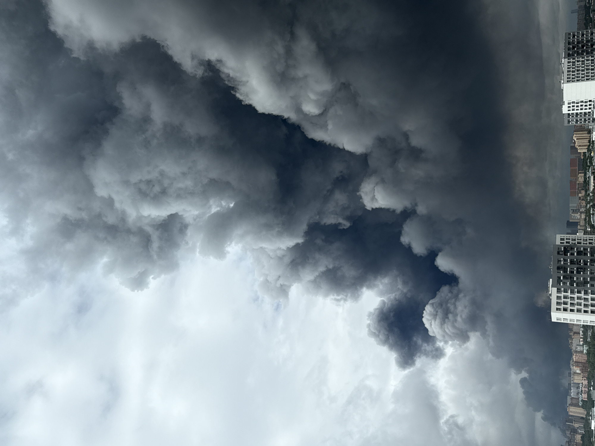 In Lyubertsy, Russia, a dramatic scene unfolds as a massive plume of thick, dark smoke billows into the sky, obscuring a significant portion of the overcast, cloudy backdrop. The smoke originates from a low-lying area, creating a stark contrast against the lighter clouds above. In the foreground, the cityscape of Lyubertsy is visible, characterized by numerous apartment buildings and other structures. A large, modern apartment building with a white facade and dark windows stands prominently in the lower right portion of the frame, suggesting a residential area. The overall impression is one of an environmental event, likely a fire, casting a pall over the urban landscape.