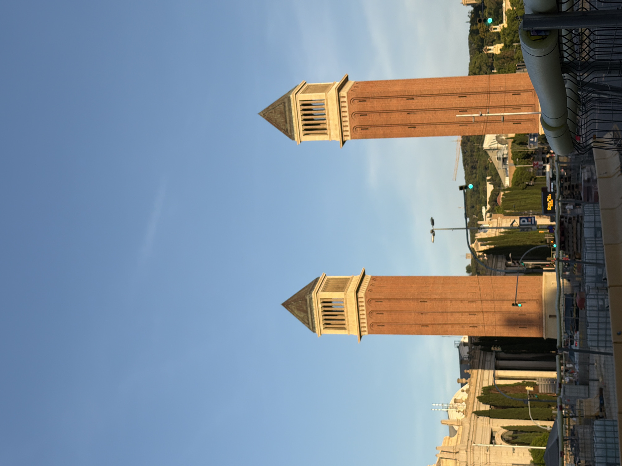 The image shows two prominent towers, likely part of the Venetian Towers in Barcelona, Spain. These towers are tall, cylindrical structures made of reddish-brown brick, topped with decorative yellow sections featuring columns and a dark pyramid-shaped roof. They stand against a clear blue sky, with the sun casting shadows. In the background, a large, neoclassical building with columns and a dome is visible, as are cypress trees and a hilly landscape covered in lush green trees. The foreground of the image depicts construction work, with scaffolding, barriers, and building materials. Traffic lights and a parking sign are also visible. The overall scene suggests a bright, sunny day in a city that blends historical architecture with ongoing development.
