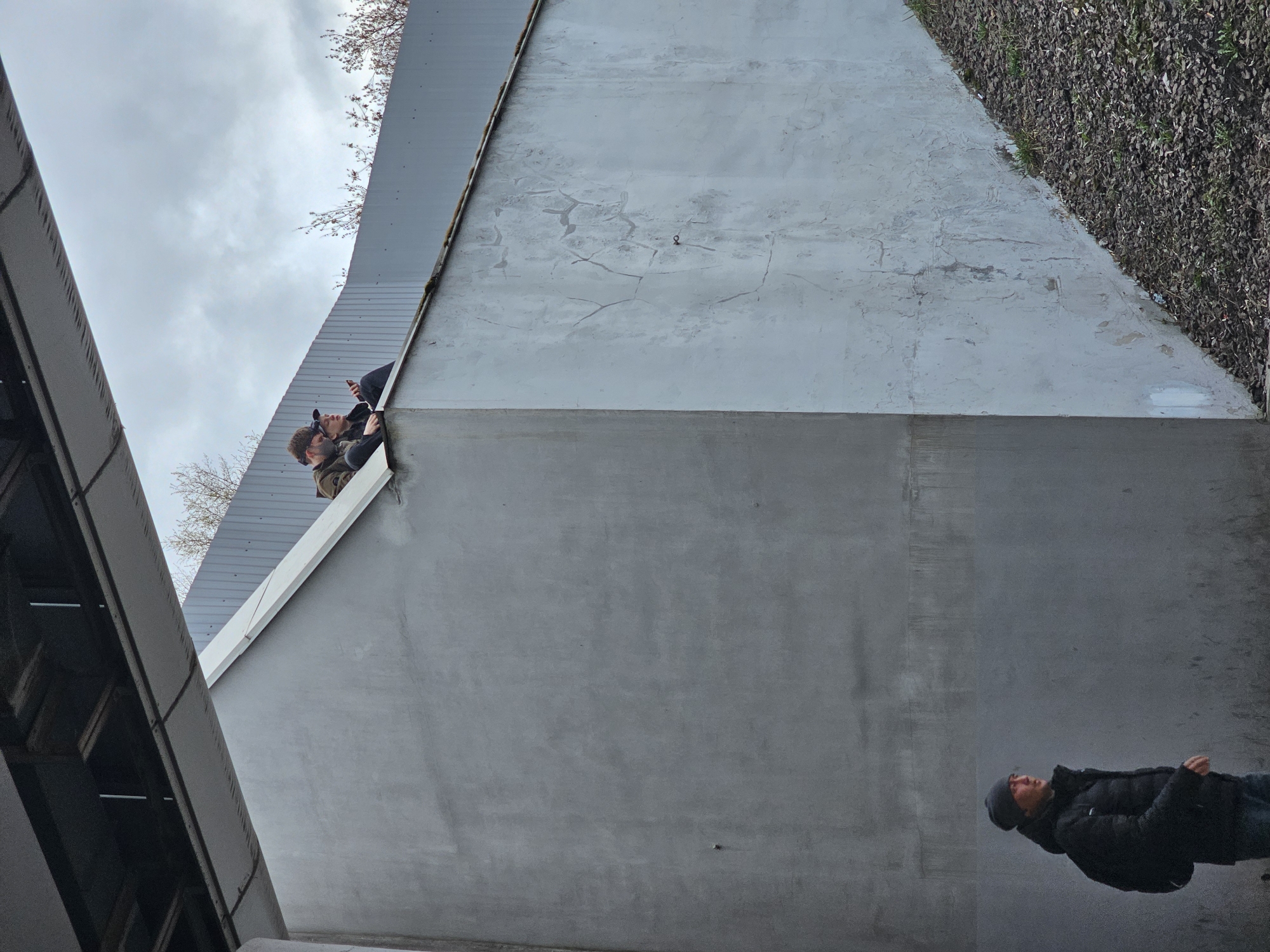 The image depicts an architectural scene in Moscow, Russia. The view is angled upwards towards a complex of gray concrete structures. Two individuals are positioned on an elevated section, possibly a bridge or walkway, observing something out of the frame. One appears to be using a mobile phone or camera. A third person, wearing a dark jacket and beanie, is standing on the ground. The environment consists of concrete walls, roof structures, and some vegetation on the upper right. The sky is overcast.