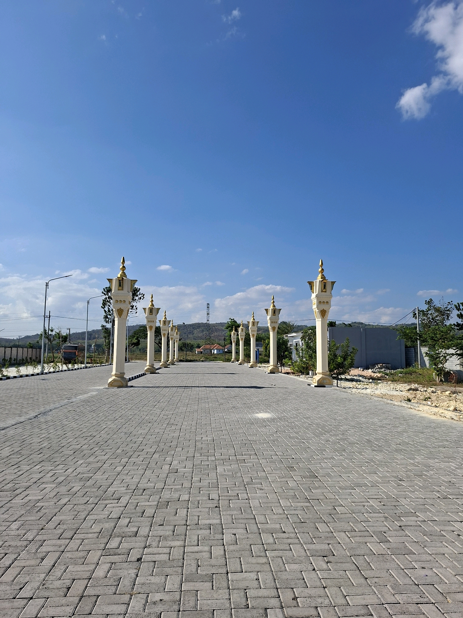 The image shows an outdoor scene in Jrengik, Indonesia, with a wide, paved walkway leading towards the horizon. The walkway is constructed of grey interlocking bricks laid in a herringbone pattern. Lining the walkway on both sides are decorative pillars, each white with gold accents, topped with a golden finial. Several pillars are visible in the foreground, and they recede into the distance, creating a sense of perspective.
The sky is a clear, bright blue with a few scattered white clouds. In the background, there are trees, some buildings, and rolling hills. To the left of the walkway, there is a streetlamp and a parked truck. The ground on the sides of the walkway appears to be unpaved, with some gravel and scattered vegetation. The overall impression is one of an open, public space under a sunny sky.
