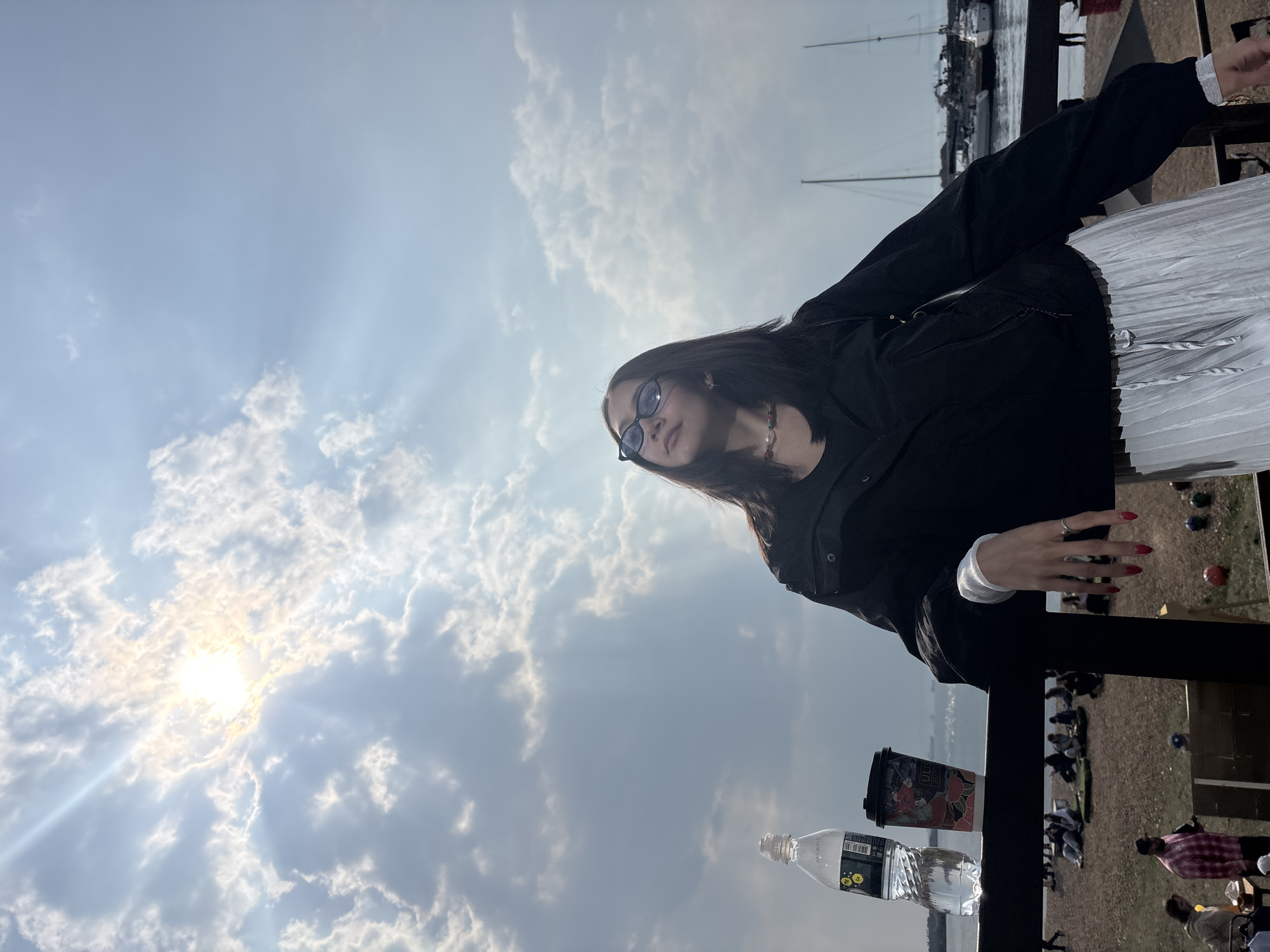 A young woman in sunglasses leans on a black railing in an outdoor setting in Vladivostok, Russia. She wears a black jacket, a white pleated skirt, and has red nail polish. Visible on the railing are a water bottle and a paper cup with Japanese-themed artwork. Behind the railing, people are seated and scattered on a beach. Boats are moored in the water in the background. The sky is partly cloudy, with the sun shining brightly.