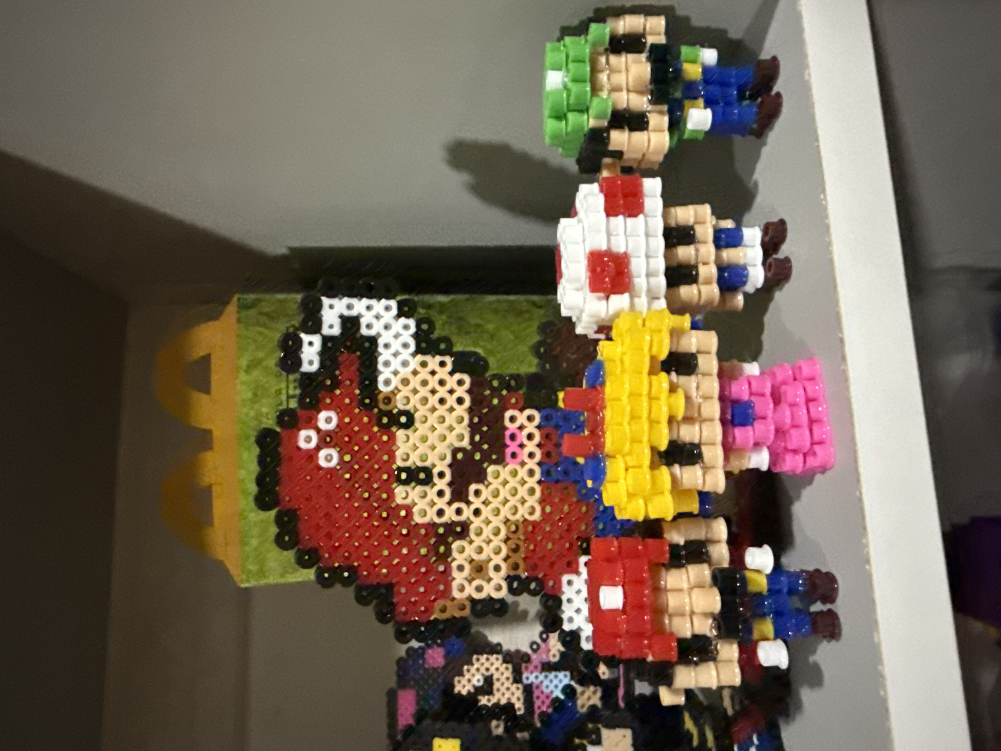 The image shows a collection of pixelated characters, presumably made of Perler beads, arranged in a stack. From top to bottom, the characters appear to be Luigi, Toad, Princess Peach, and Mario. The background includes a yellowish object with handle-like features, possibly a plastic container, and a section of what appears to be a shelf or cabinet interior with white walls. There is no evidence to determine the location in the city of Puebla, Mexico.