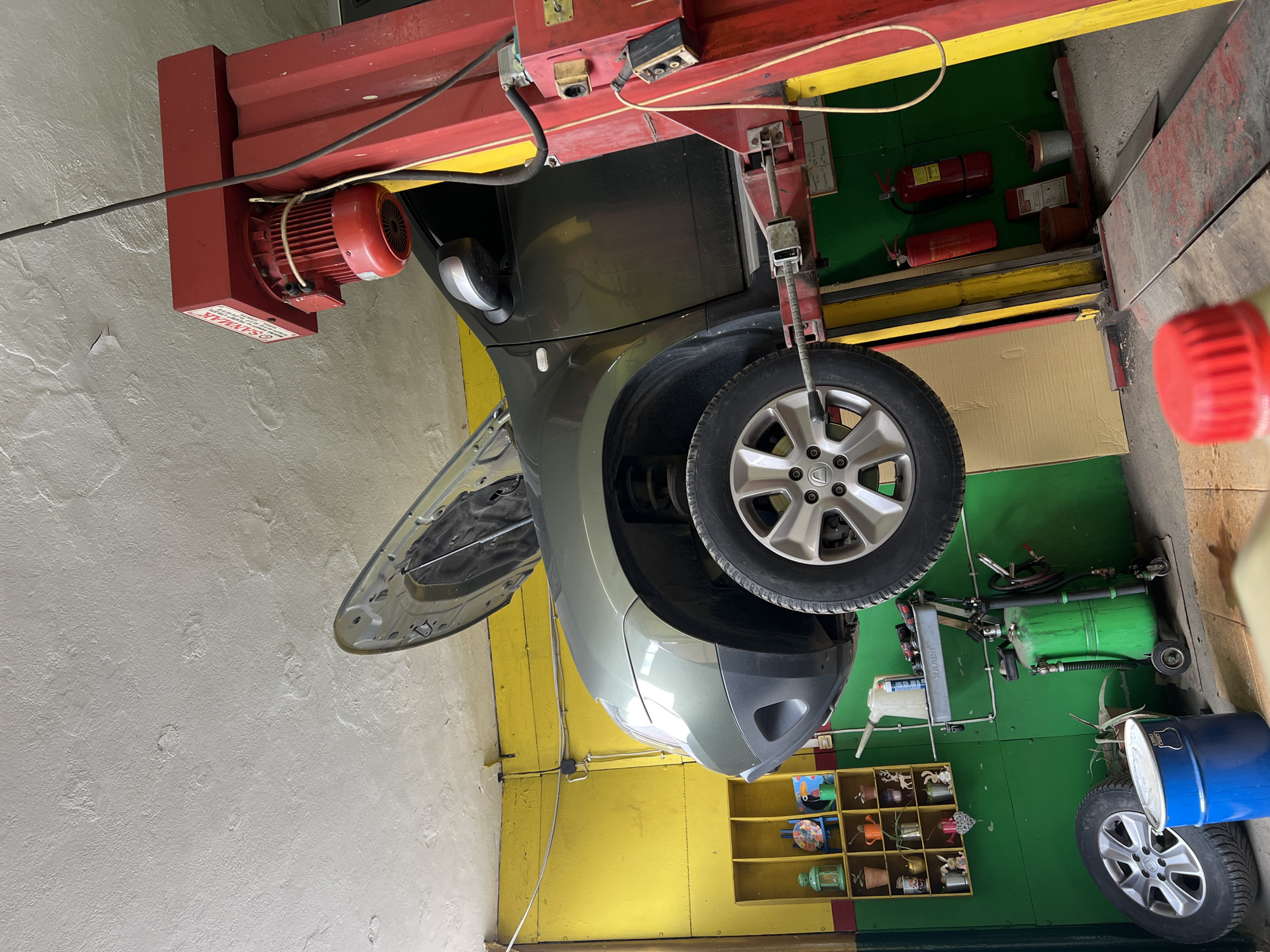 A car, a grey Dacia, is elevated on a red and yellow mechanical lift within a garage setting. The front driver-side door is open, revealing the interior. A tire with silver rims and the Dacia logo is prominent. The walls are painted yellow and green. Tools, a green air tank, and a blue barrel are on the ground. Fire extinguishers are mounted on the wall, with a red cap nearby. A red motor is fixed to the lift. Shelves containing decorative items are also visible. The location may be Plovdiv, Bulgaria. No people are visible.