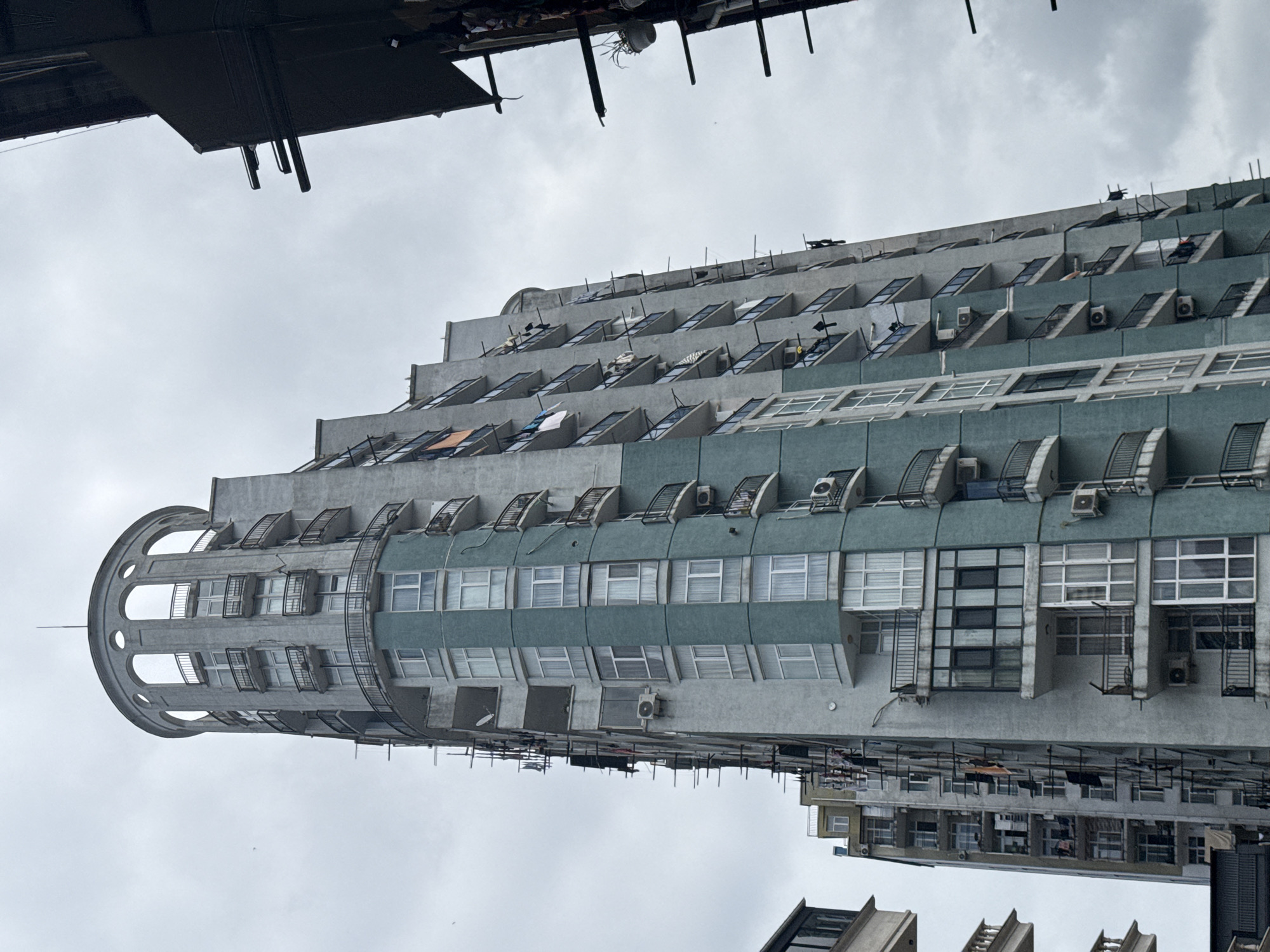 A low-angle, full-shot photograph shows the exterior of a tall, multi-story residential building in an urban environment. The building is comprised of numerous windows and balconies, with some balconies appearing to have been enclosed with glass. The facade of the building is a combination of light gray and teal green panels. Clothes are visible hanging from some of the balconies, suggesting that residents are drying laundry. The building has a distinctive rounded top with arched openings and a railing. The sky is overcast and gray. Another building is partially visible on the right side of the frame, and a portion of another building is visible on the left. The overall impression is of a dense urban setting with a focus on residential architecture.