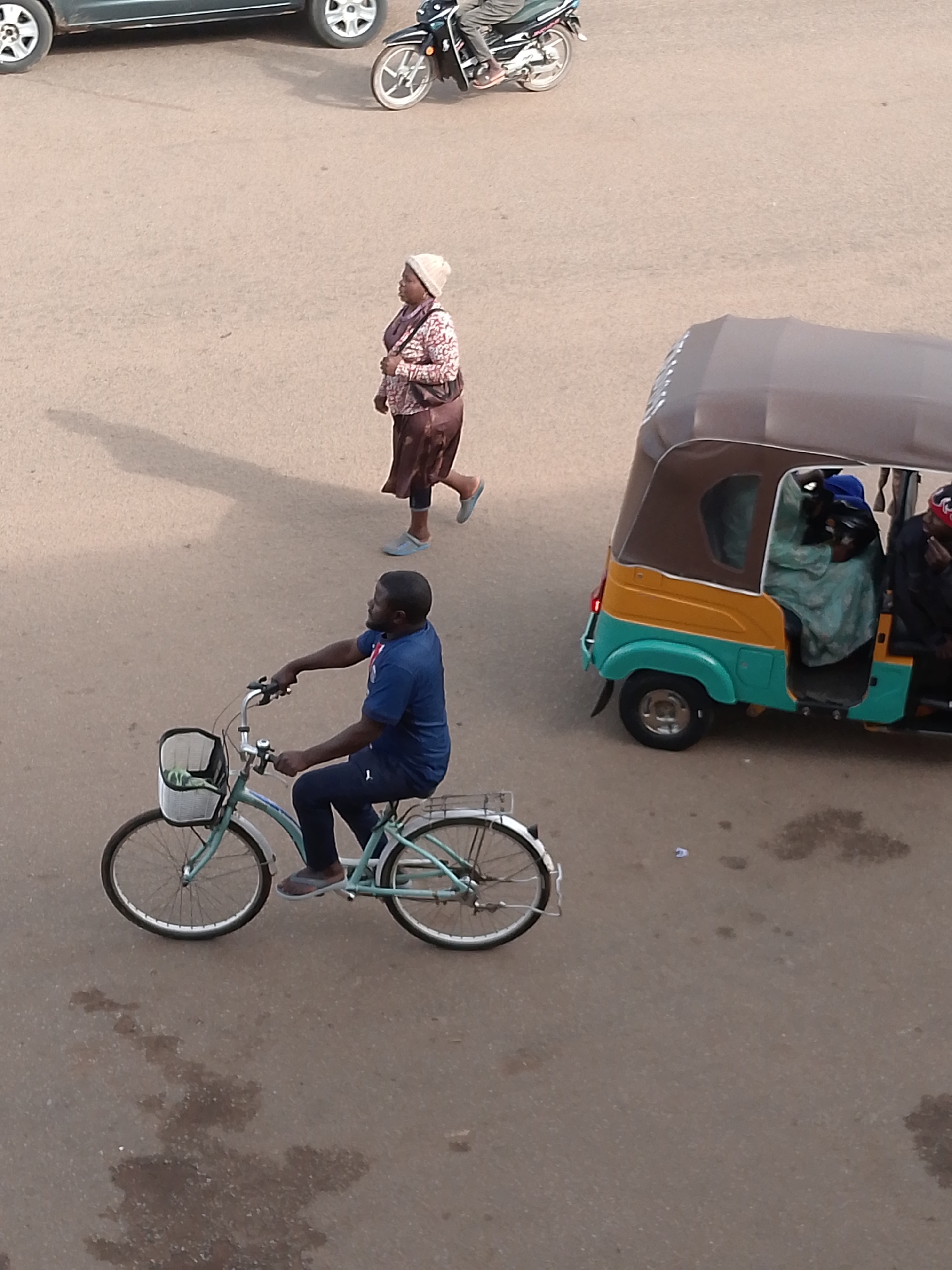 In Bukuru, Nigeria, a man rides a light blue bicycle with a basket on the front. He is wearing a dark blue t-shirt and dark pants. To the right, a colorful auto-rickshaw, with a brown canopy and yellow and turquoise body, transports at least three passengers. One passenger in the front is wearing a blue headscarf and a light green garment, possibly a hijab and robe. Another passenger in the back is wearing a red and black patterned head covering. Further in the background, a woman in a patterned top, brown skirt, and a light-colored head covering walks away from the viewer. Behind her, a motorcycle is parked. The scene takes place on a paved surface, likely a road or open area, with some dark patches indicating stains or wet areas.