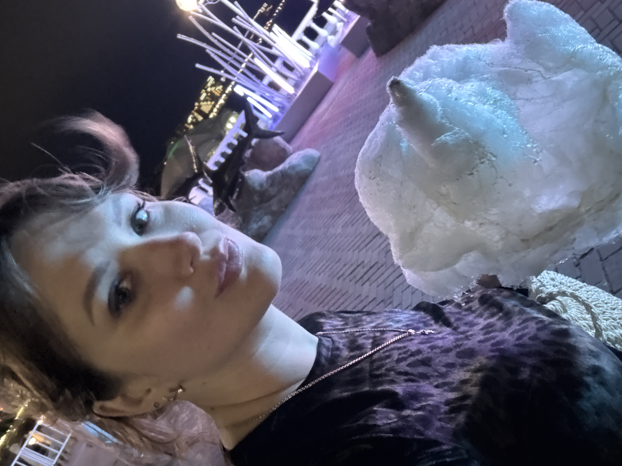 The image shows a woman in Gelendzhik, Russia, taking a selfie at night. The background includes illuminated architectural details and a sculpture. The woman is wearing a dark animal-print top and a necklace. She is holding a large white object, possibly cotton candy or a similar confection.