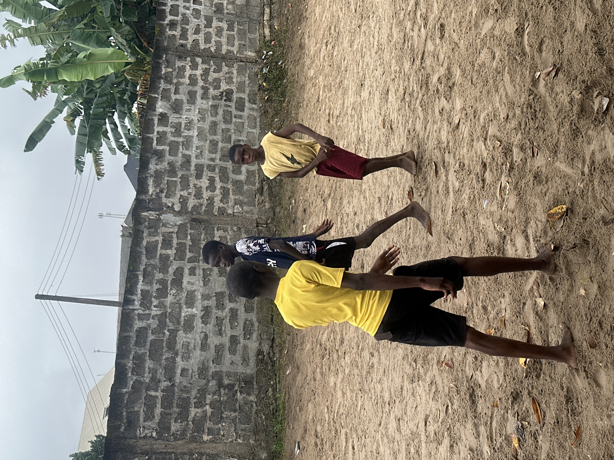 Three young boys are standing on a sandy patch in a somewhat dilapidated urban area, possibly Badagry, Nigeria. The boy in the foreground on the left is wearing a yellow t-shirt and black shorts, with his back mostly to the camera. To his right, a boy in a dark blue t-shirt with a white pattern and black shorts is walking away, also with his back to the camera. The third boy, on the right, is wearing a yellow sleeveless shirt with a graphic design and maroon shorts. He is facing the camera, looking directly at it with a neutral expression. Behind the boys is a rough, grey brick wall, partially covered with vegetation and debris. In the background, there is a utility pole with electrical wires stretching across the cloudy sky, and a glimpse of buildings with light-colored roofs. Banana trees are visible to the right of the frame, suggesting a tropical climate. The overall scene depicts a casual, outdoor gathering of children in a modest urban environment.