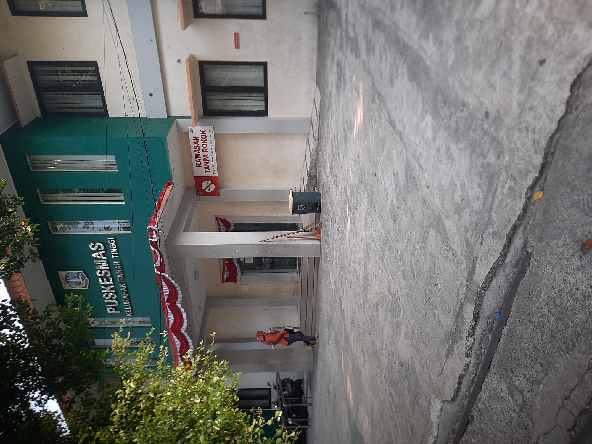 The image shows the exterior of the Puskesmas Kelurahan Tanah Tinggi, a public health center located in Central Jakarta, Indonesia. The building has a green facade on the upper story with the name of the facility prominently displayed. Below the green section, a white awning is decorated with red and white bunting. A "No Smoking" sign is visible on the right side of the entrance. In front of the building, a concrete walkway leads to the entrance. To the left of the entrance, a person wearing an orange headscarf and an orange top is walking away from the building. A broom and a trash bin are positioned near the entrance. The ground in front of the building is made of cracked concrete. Lush green trees partially obscure the view of the building from the left side of the frame. Motorcycles are parked in the background. The lighting suggests it is daytime, with sunlight casting shadows.