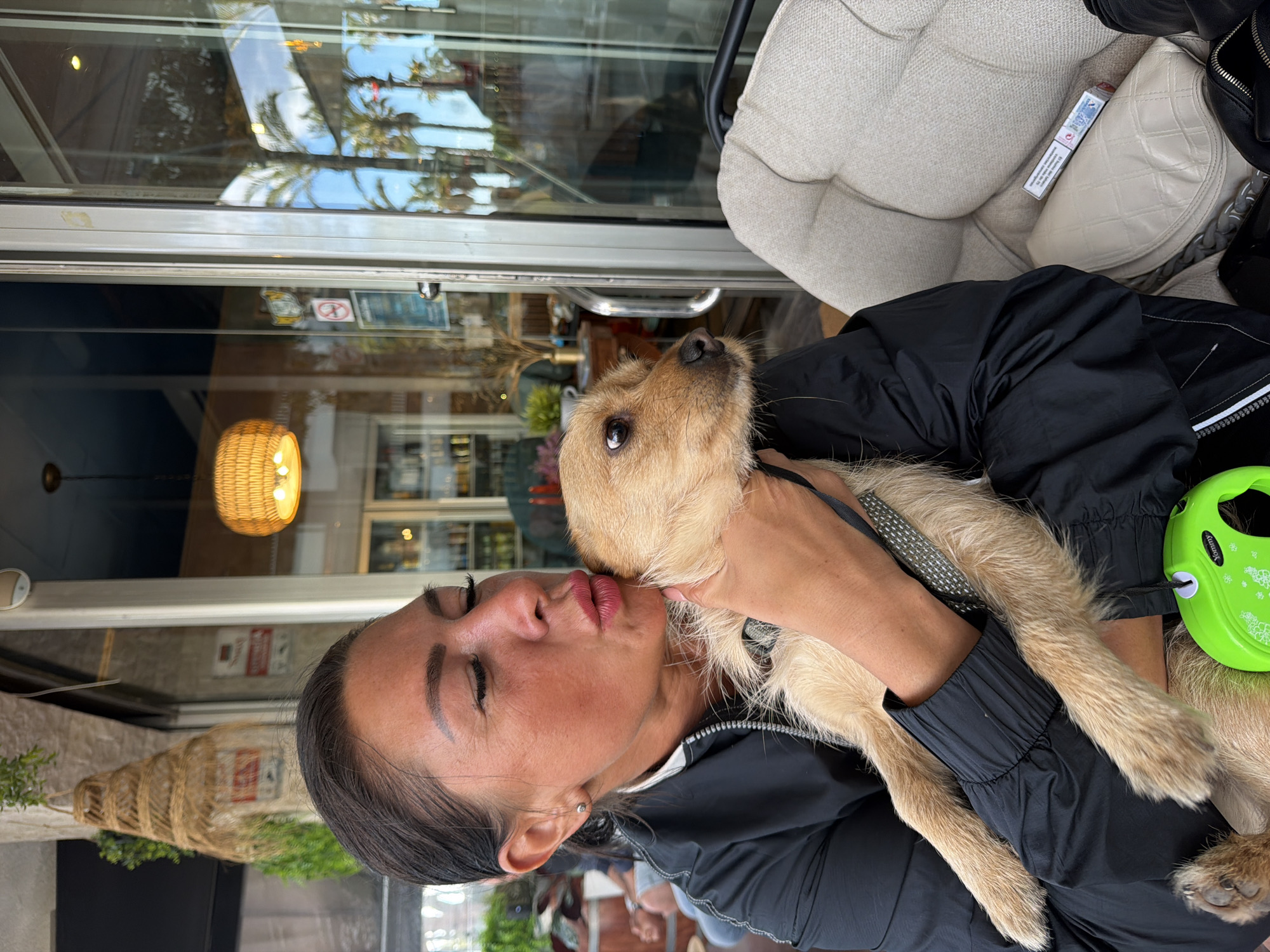 A person with dark hair is kissing a light-brown, medium-sized dog, appearing to be a terrier mix. The person is wearing a black jacket. The dog is wearing a black collar with a gray leash and is held in the person's arms. A green retractable leash is visible next to the dog on the person’s lap. Behind the person and the dog is a glass storefront, reflecting palm trees and buildings, suggesting an outdoor location in Salou, Spain. A wicker light fixture is visible inside the storefront. A patterned handbag and a pack of cigarettes are visible on the seat behind the person.