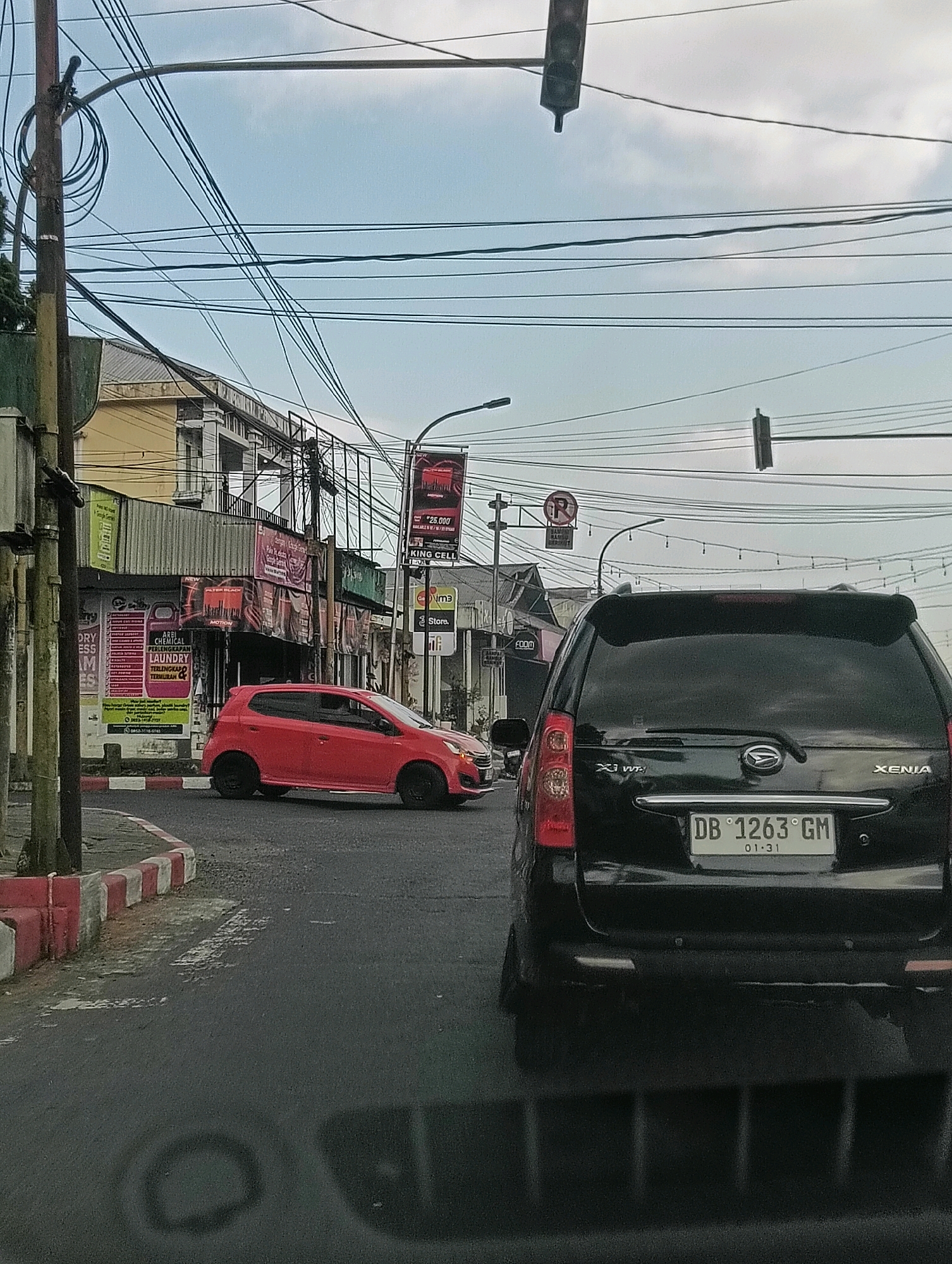 The image depicts a street scene in Tomohon, Indonesia, under an overcast sky. In the foreground, a black SUV is stationary, facing away from the camera. Its license plate reads "DB 1263 GM 01-31". To the left of the SUV, a bright red hatchback is turning at an intersection. The street is paved, with a red and white striped curb visible on the left side. Various commercial buildings line the street, displaying signage for businesses such as "KING CELL", "Store", and what appears to be a laundry service. A complex network of overhead power lines and traffic lights crisscrosses the sky, typical of urban infrastructure. The overall impression is that of a typical urban traffic environment in Indonesia.