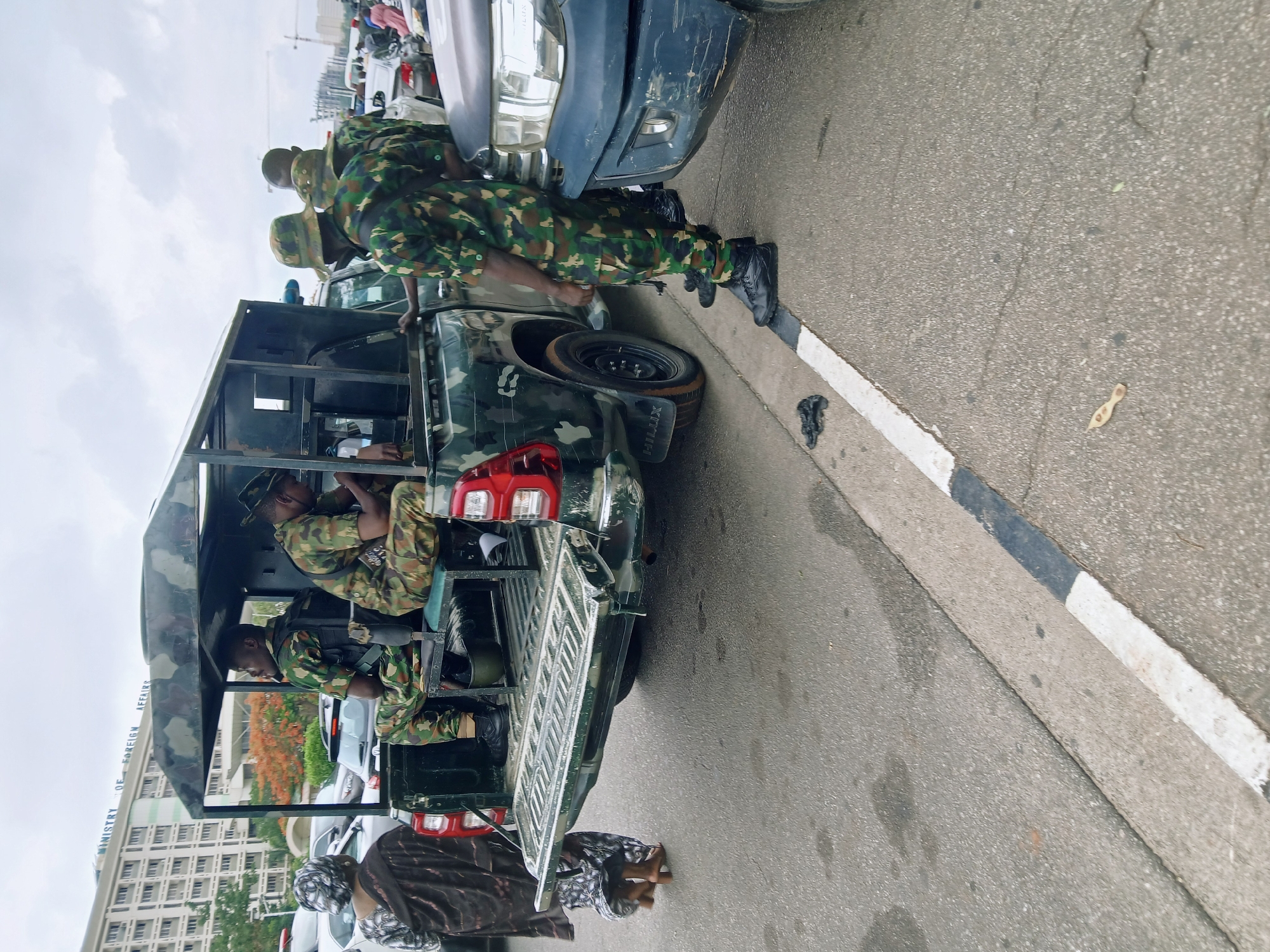 The image shows a scene in Abuja, Nigeria, in front of the Ministry of Foreign Affairs building. A military vehicle, camouflaged in green and brown, is parked on the side of the road. At least three uniformed soldiers are either inside or near the vehicle. One soldier stands near the front of the vehicle, while two are in the back, visible through the open tailgate. Two individuals, possibly women, are kneeling on the ground near the vehicle's rear. Other civilian vehicles are visible in the background, suggesting a busy urban environment. The road has painted lines and appears to be asphalt.