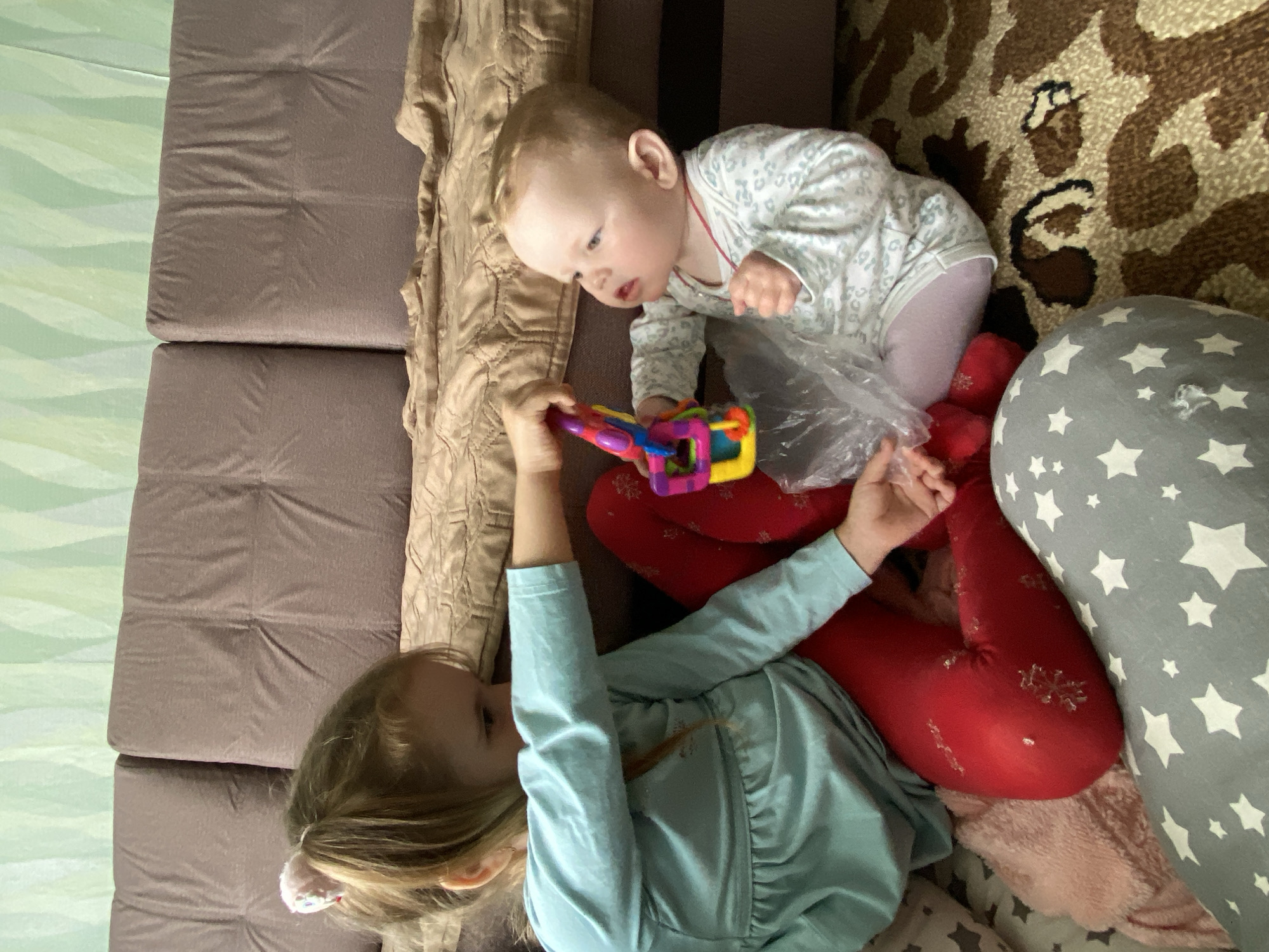 This is an overhead, slightly angled photograph depicting two young children playing indoors in Фоки, Russia. An older child, with light blonde hair, is lying on her back on a brown upholstered couch. She is wearing a light blue long-sleeved top and red leggings adorned with white snowflake patterns. Her head is towards the left of the frame, and she holds a colorful, multi-piece plastic toy, possibly a rattle or teether, in her hands.
Adjacent to her, a baby with light blonde hair is on her knees, leaning forward and looking towards the toy held by the older child. The baby is wearing a white long-sleeved onesie with light grey patterns and is holding a clear plastic bag in her hands. The children appear to be engaged in a moment of shared play or interaction, with the baby showing curiosity towards the toy.
The setting is a cozy indoor space, likely a living room. The children are positioned on a brown upholstered couch or sofa, partially covered by a beige textured throw blanket. To the right of the children, a patterned rug or carpet with brown, beige, and black abstract or animal print designs is visible. A grey pillow with white star patterns is near the baby. In the background to the left, a light green wall or wallpaper with a subtle wavy pattern can be seen. A pink blanket or soft fabric is also partially visible underneath the older child's legs. The general lighting suggests it is daytime. No visible text is present in the image.