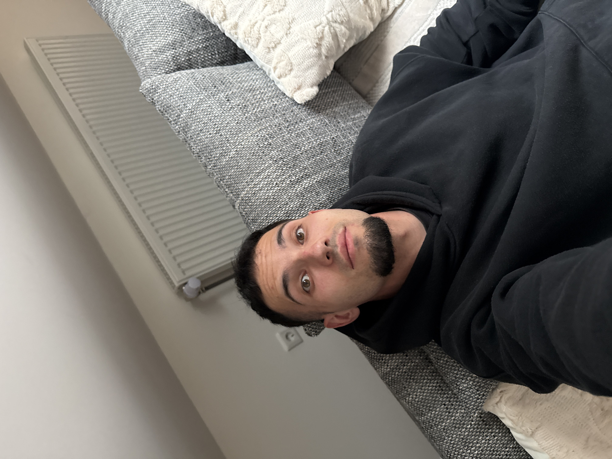 A man with short dark hair, a goatee, and light brown eyes is lying on a grey couch, looking at the camera. He is wearing a black hoodie. The couch has textured grey cushions and one cream-colored cushion with a textured pattern. In the background, a white radiator is visible on a light-colored wall, and a light switch is mounted on the wall. The overall environment appears to be indoors, likely a living room. The image is cropped in a way that suggests it's a selfie. Garmisch-Partenkirchen, Germany is a town known for its alpine scenery and winter sports, but this indoor scene does not provide specific visual cues to confirm its location within the town.