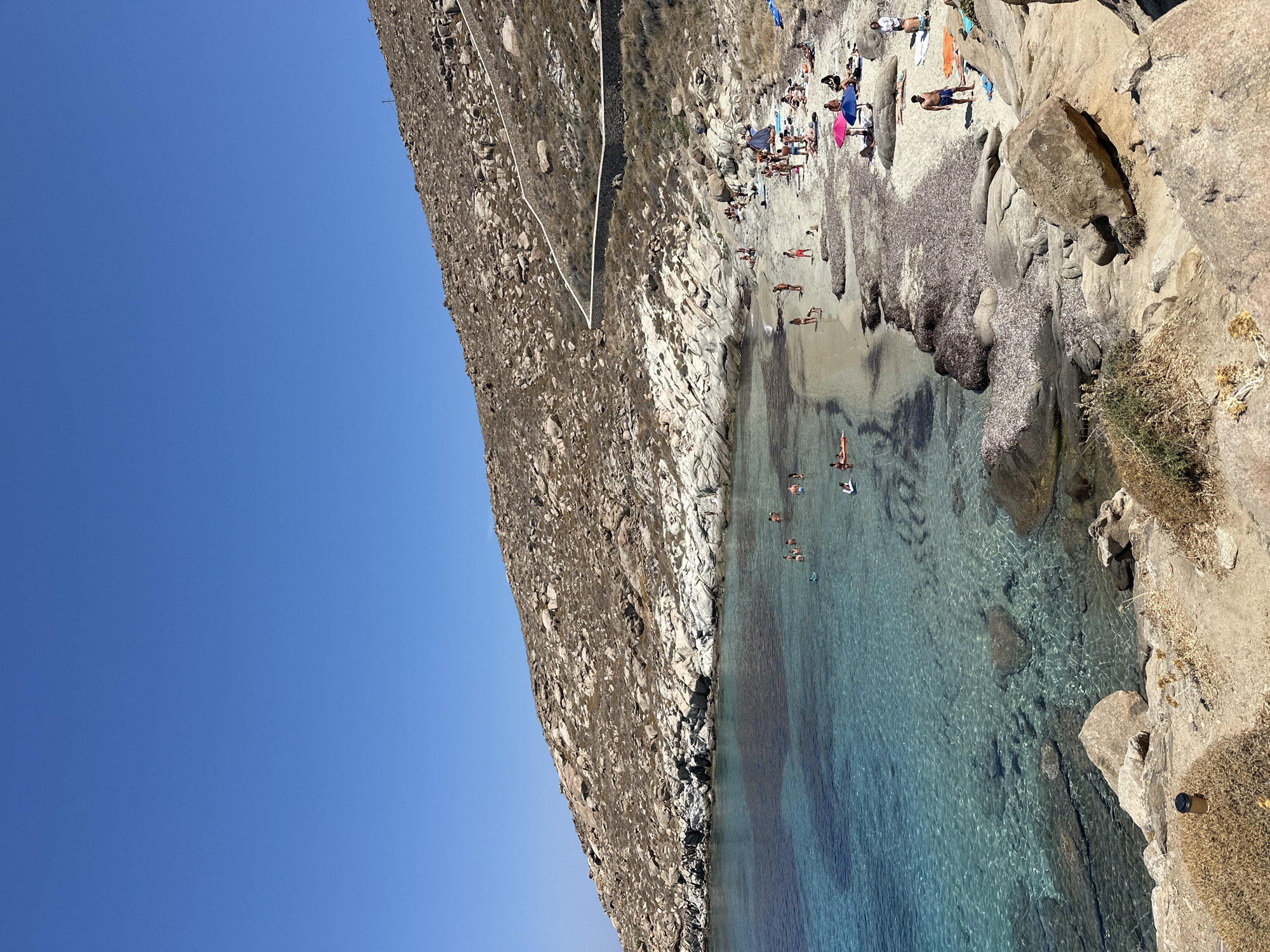 The image depicts a scenic view of a beach in Agios Ioannis Diakoftis, Greece. A clear blue sky dominates the upper portion of the frame, transitioning into a rocky, arid hillside that slopes down towards the sea. The hillside is dotted with numerous light-colored rocks and sparse vegetation. A stone retaining wall winds its way along the contour of the hill.
The coastline features a sandy beach with scattered rocks and patches of dried seaweed. The sea is crystal clear, revealing the sandy bottom and submerged rocks in its turquoise waters. Several people are visible enjoying the beach and the water. Some are swimming or wading in the sea, while others are sunbathing on towels and beach chairs on the shore. Beach umbrellas in pink and blue provide shade for some of the beachgoers. A few individuals are standing or walking along the beach. In the foreground, large boulders and dry brush frame the scene, giving a sense of the natural, rugged terrain.