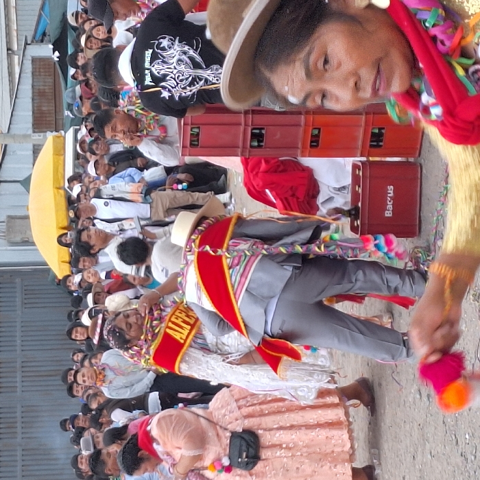 A lively outdoor scene features a crowd of people gathered for what appears to be a festival or celebration. Many are dressed in traditional clothing and hats. Colorful streamers, possibly confetti, hang in the air, adding to the festive atmosphere. Individuals stand on top of cases that say "Bacus". The focus is on a group of participants in the foreground, with a woman in a red sash and another in light-colored attire adorned with a red banner, suggesting they might be involved in a parade or procession.