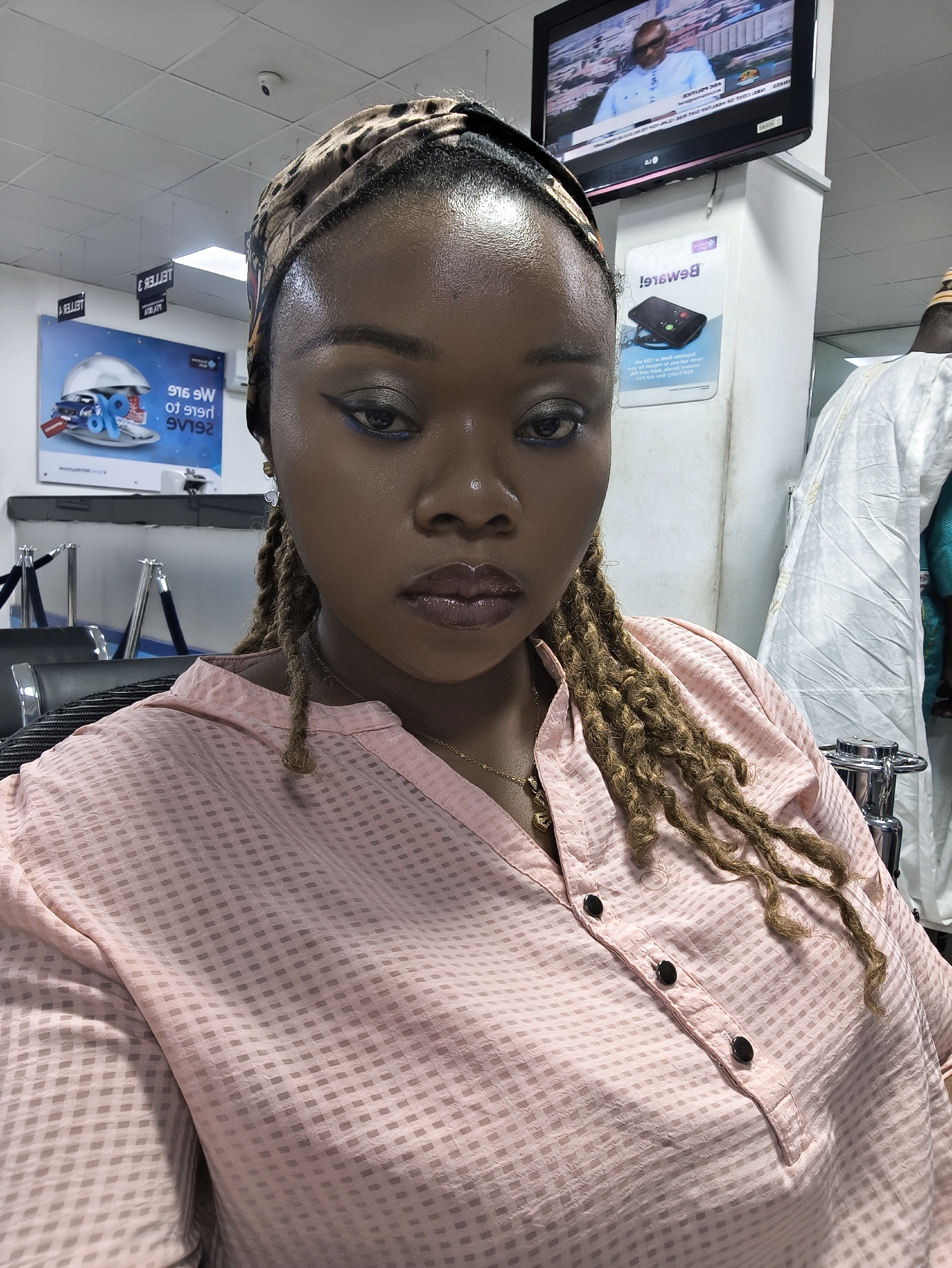 A close-up, eye-level portrait shows a woman in what appears to be an indoor public space, possibly a bank or office waiting area. The woman, with dark skin, looks directly at the camera with a neutral, serious expression. She wears a patterned headband, possibly with leopard print, over her dark hair, which is styled into long, light brown dreadlocks visible cascading over her right shoulder and chest. She has prominent makeup, including shimmery grey eyeshadow, bold blue eyeliner, and glossy dark pink or plum lipstick. Gold-toned, heart-shaped earrings are visible. Her top is a light pink, long-sleeved collared shirt with a subtle checkered pattern and three visible dark buttons down the placket, over which a delicate gold chain necklace with a small pendant rests.
The background is an indoor setting with white walls and a white drop ceiling. A security camera is mounted on the ceiling. On the left, a large poster is visible, showing a silver cloche covering what appear to be gift boxes and a small car, with a large percentage symbol. Mirrored text on this poster reads "916 W TO 9192" and "SVELTA". Below it, a black queuing stanchion with a black rope is partially visible.
To the right, a television screen is mounted high on a wall, displaying a man in a light-colored outfit, likely a news anchor or guest, with some mirrored text on the screen, indicating a broadcast. Below the TV, another poster features a smartphone on a stand and the mirrored word "Beware!" at the top. Further right, a person in a white, traditional garment (possibly an agbada or similar attire) is partially visible. The overall lighting suggests artificial indoor illumination.