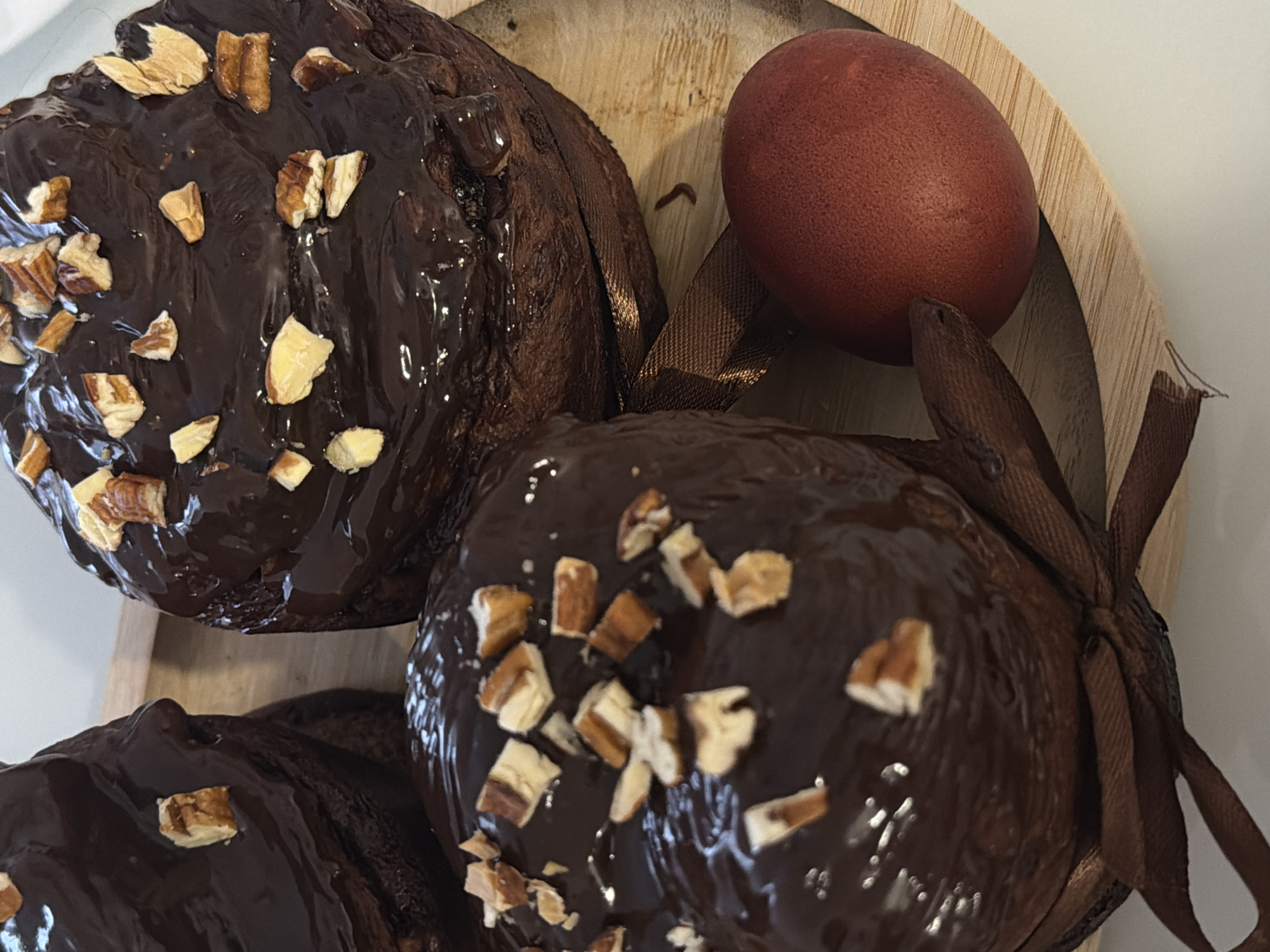 The image shows a wooden platter containing three chocolate-glazed cakes sprinkled with chopped nuts and an egg dyed reddish-brown. A brown ribbon is tied around one of the cakes. The background is a light-colored surface. There is no indication of the city of Khimki, Russia in the image.