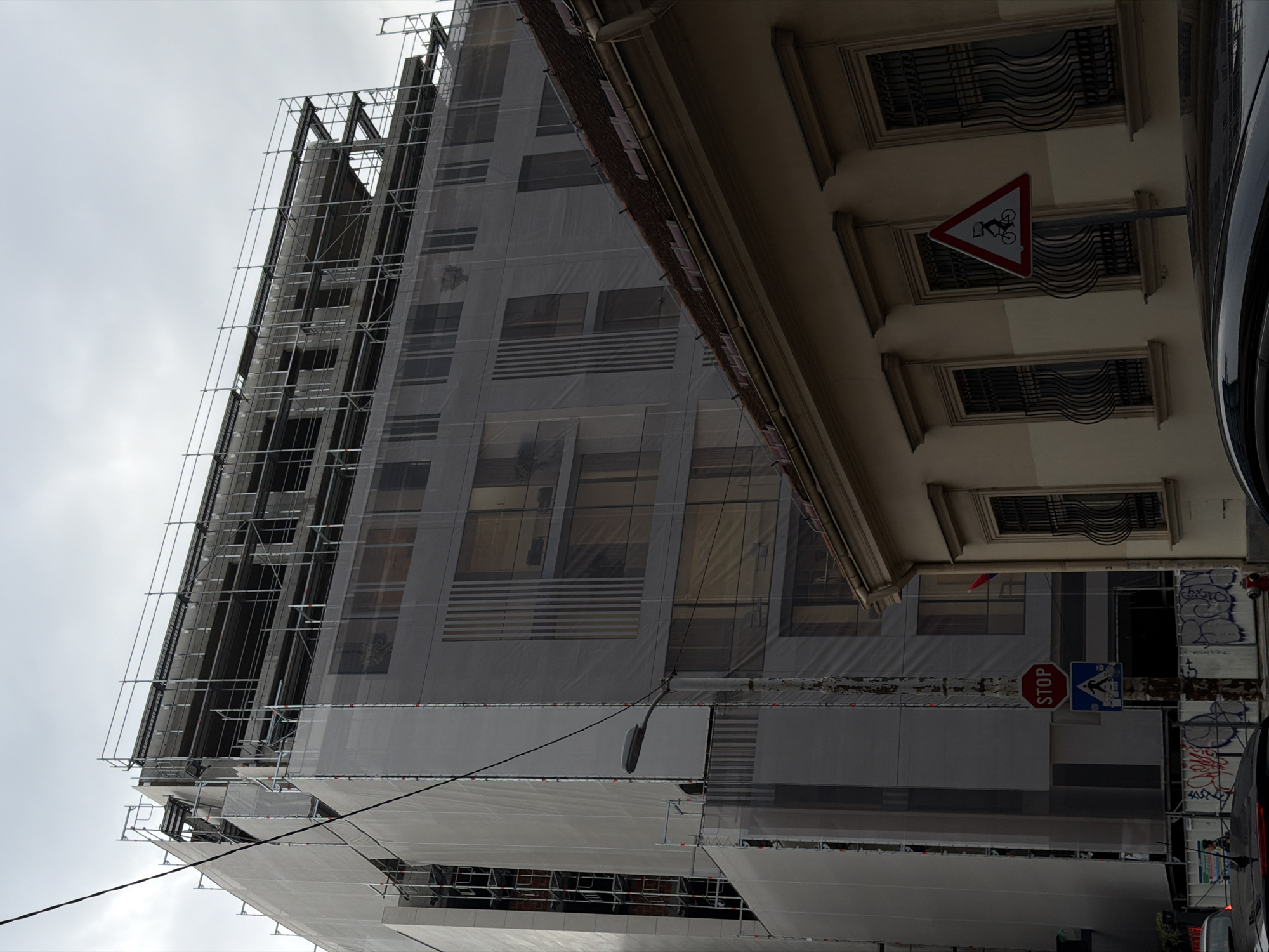 The image shows a street scene in Belgrade, Serbia, with a large building under construction covered in scaffolding and white sheeting. A smaller, older building with a tiled roof and barred windows is in the foreground, partially obscuring the construction site. A stop sign and a pedestrian crossing sign are visible on a pole in front of the building. A lamppost is also present. The sky is overcast. The street appears to be a public area with some graffiti on a temporary fence. The construction site suggests ongoing urban development.