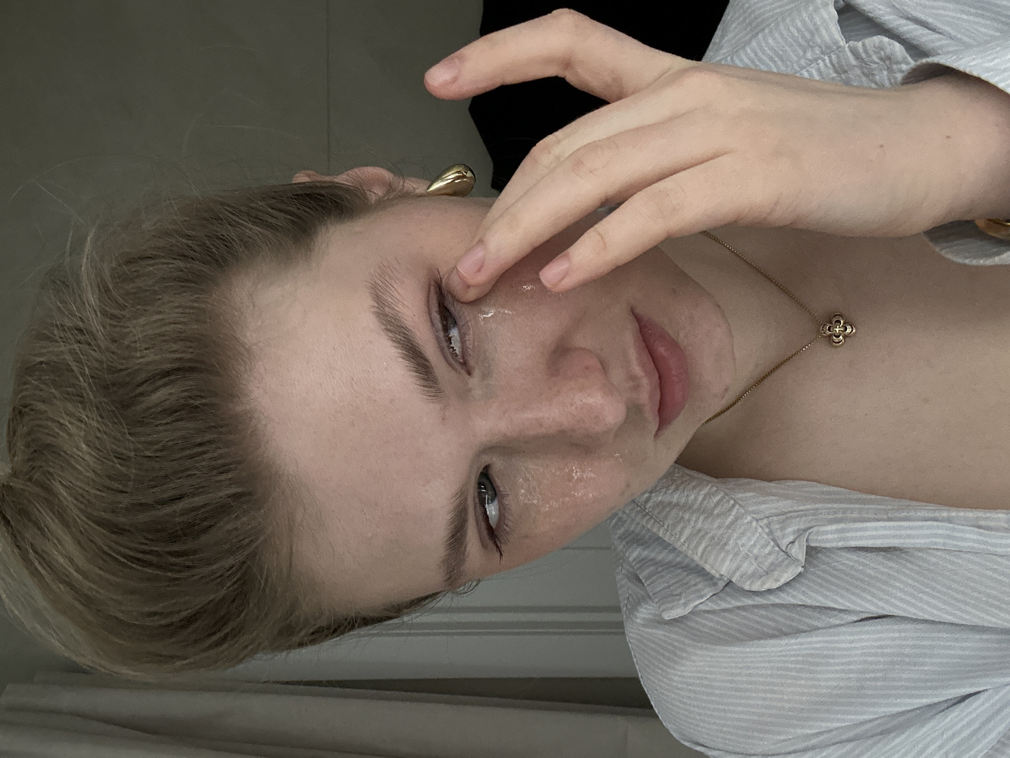 A young woman with light blonde hair is depicted indoors. She is wearing a light blue and white striped collared shirt and a gold necklace with a clover pendant. Her right hand is near her face, with her fingers positioned near her right eye. She appears to have moisture on her face, possibly tears or a cosmetic product. A gold earring is visible on her left ear. The background shows a white wall and possibly part of a bed. The image is taken from a low angle and rotated counterclockwise. There is no evidence to determine that the location is in the city of Kazan, Russia.