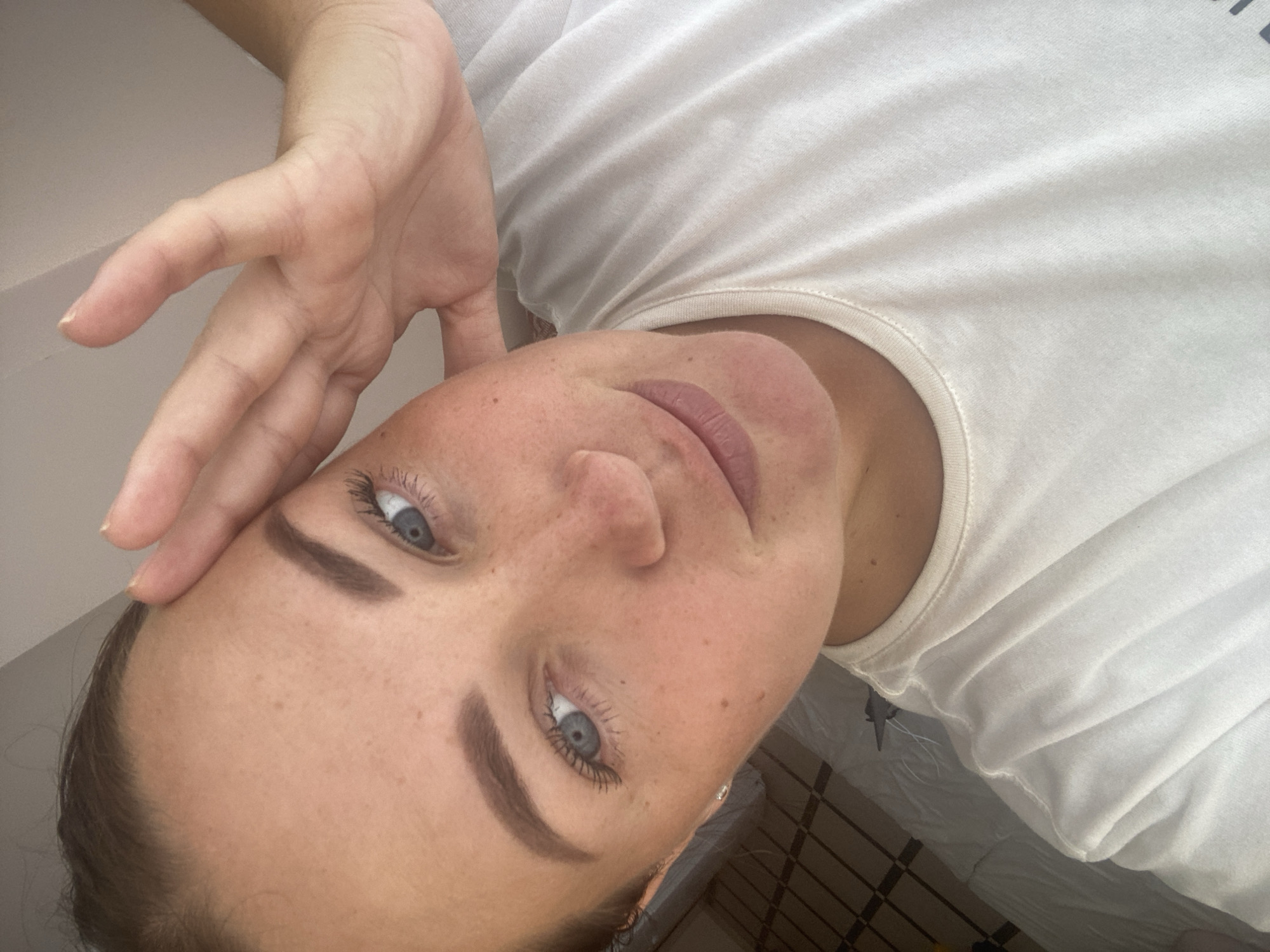 A woman is taking a selfie with her right hand on her forehead and her left eye is partially visible. She is wearing a white t-shirt. Her skin is fair with freckles. Her eyebrows are dark and well-defined, and her eyelashes are long. The background is blurred and appears to be an indoor setting, possibly a bedroom with a bed and a wooden floor. There is no indication of the city of Sochi, Russia in the image.