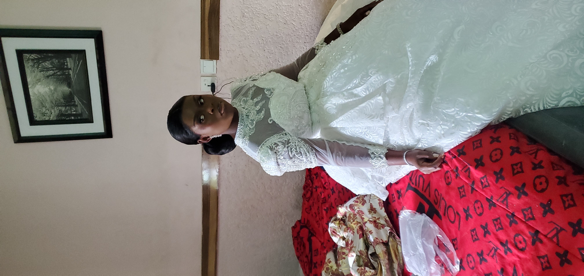 A young Black woman in a white wedding dress is seated on a bed with a red Louis Vuitton patterned blanket. She is looking directly at the camera. Her dark hair is pulled back, and she is wearing jewelry. The room has light pink walls. A framed black and white picture hangs on the wall behind her. A light switch is visible near a wooden trim. The location is Udu, Nigeria.