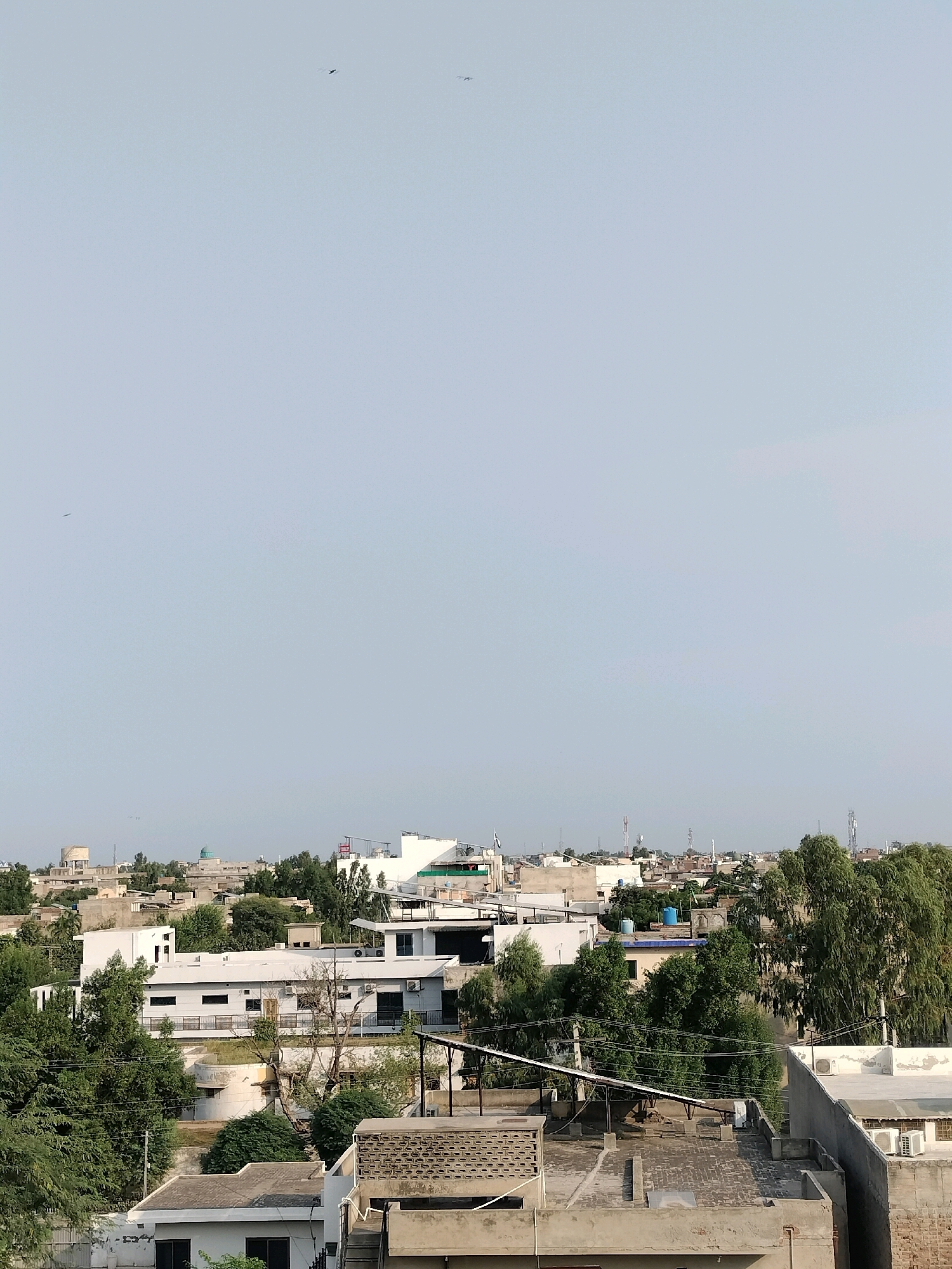 This image captures a panoramic view of a densely populated urban area, likely within Sargodha, Pakistan, under a pale, hazy sky. The foreground is dominated by rooftops of various buildings, some with exposed concrete, others with tiled surfaces, and a few with visible water tanks. Lush green trees are interspersed throughout the landscape, many growing tall and full, suggesting a warm climate.
In the middle ground, a cluster of multi-story buildings, primarily white, with visible windows, air conditioning units, and some darker architectural features, form a more organized section of the cityscape. The presence of telecommunication towers and satellite dishes indicates a modern, connected environment. Further in the distance, the urban sprawl continues, with numerous buildings of varying heights and architectural styles stretching towards the horizon. A prominent blue water tank is visible on top of one of the buildings.
The sky is a uniform, light blue with a slight greyish tint, indicating either a hazy atmosphere or the early stages of a less intense day. Two small, dark shapes resembling birds or insects are visible in the upper portion of the sky, adding a sense of scale and dynamism to the otherwise static cityscape. No people are clearly discernible in the image. The overall impression is one of a thriving, somewhat informal, urban environment within a Pakistani city.
