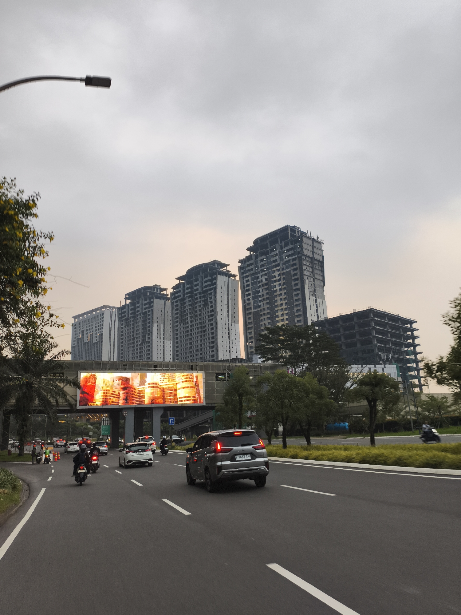 This vertical shot captures a busy street scene in Babakan Madang, Indonesia, during what appears to be late afternoon or early evening under an overcast sky. The foreground features a multi-lane asphalt road with clear white markings, populated by a stream of traffic including several cars and numerous motorcycles. A gray SUV with the license plate "F 1663 AAV" is prominently visible in the right lane, driving away from the viewer.
To the left, a median strip with dense green foliage separates the road, where a few people on motorcycles are paused. A curved street light extends into the top-left of the frame. An elevated structure spans across the road, featuring a large, brightly illuminated digital billboard displaying a vibrant advertisement with warm orange and yellow hues depicting stylized cityscapes.
In the midground and background, various trees, including palm trees and other dense green foliage, line the road. Behind these, several modern high-rise buildings dominate the skyline. Some of these structures appear completed, while others are clearly under construction, showing exposed concrete frames and unfinished sections. A small blue sign with a white U-turn arrow is visible beneath the elevated platform. The scene depicts a dynamic urban or developing area with ongoing infrastructure and building growth.