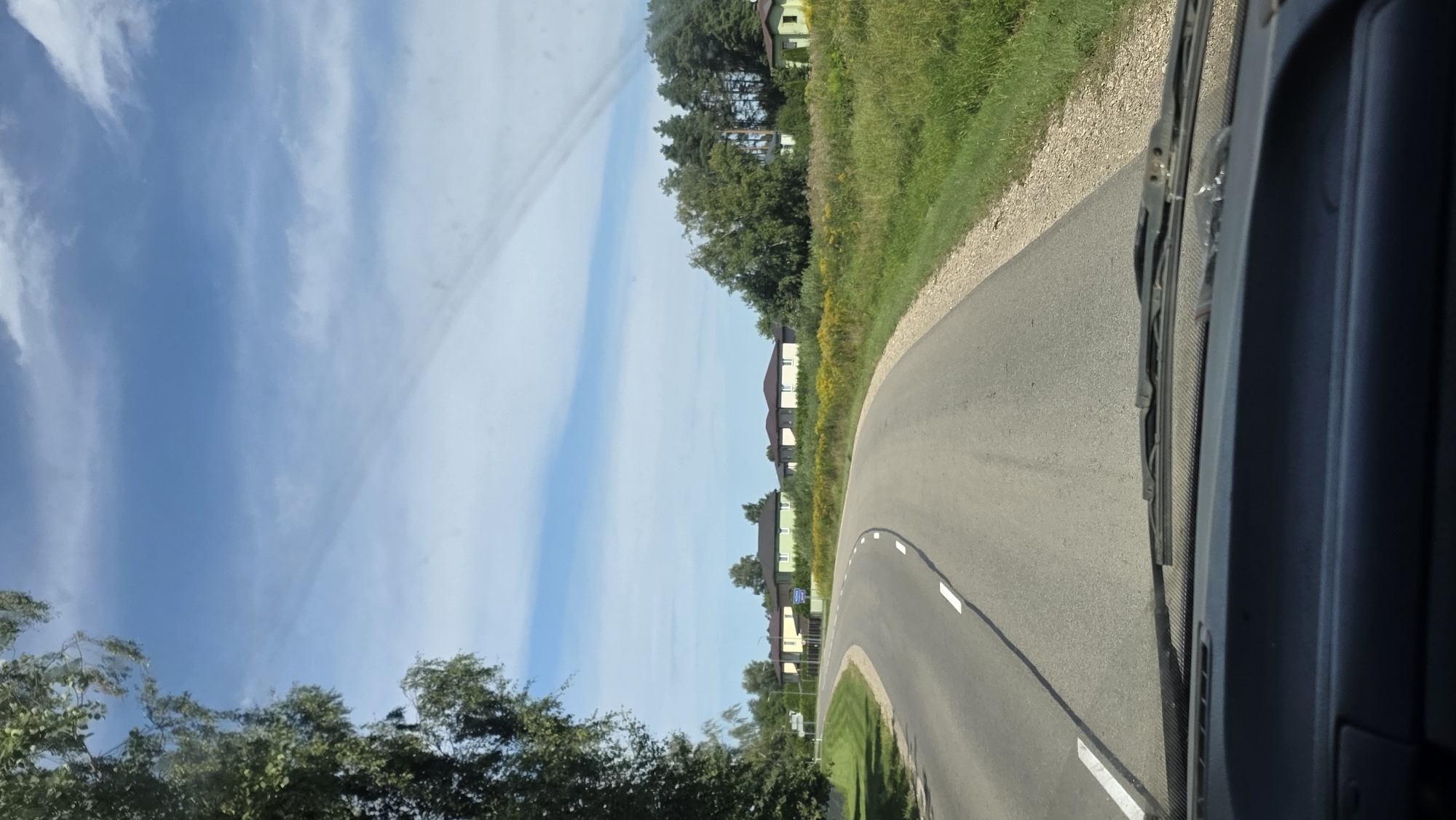A curved asphalt road with white lane markings passes through a rural area with green fields and trees under a partly cloudy blue sky. Several houses with dark roofs are visible on the right side of the road, situated amongst trees and vegetation. The image is taken from the perspective of someone inside a vehicle, looking out through the windshield, which has windshield wipers visible at the bottom. The setting appears to be a daytime scene in a village or small town environment.