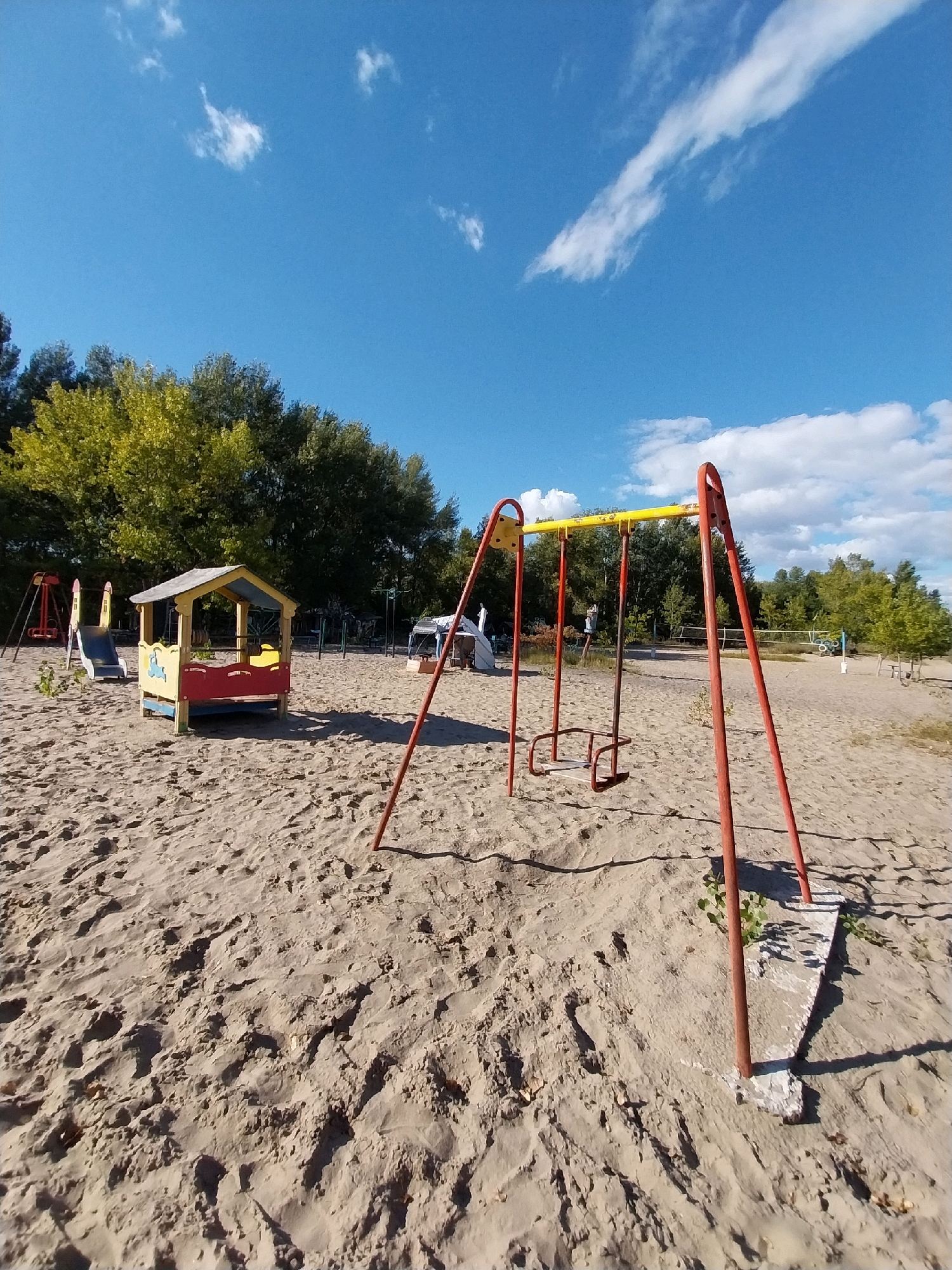 The image depicts a deserted sandy playground in Dnipro, Ukraine, on a clear, sunny day. The playground features a red and yellow swing set in the foreground, with two empty swings. Behind it, a small, colorful playhouse with a red and yellow roof sits on the sand. A blue slide is attached to a red frame to the left of the playhouse. Trees with green and yellow foliage form a backdrop, with a bright blue sky and scattered white clouds above. The sandy ground is marked with footprints and shadows cast by the playground equipment and trees. In the distance, other playground structures and trees are visible, along with what appears to be a volleyball net. The scene is tranquil and devoid of human activity.