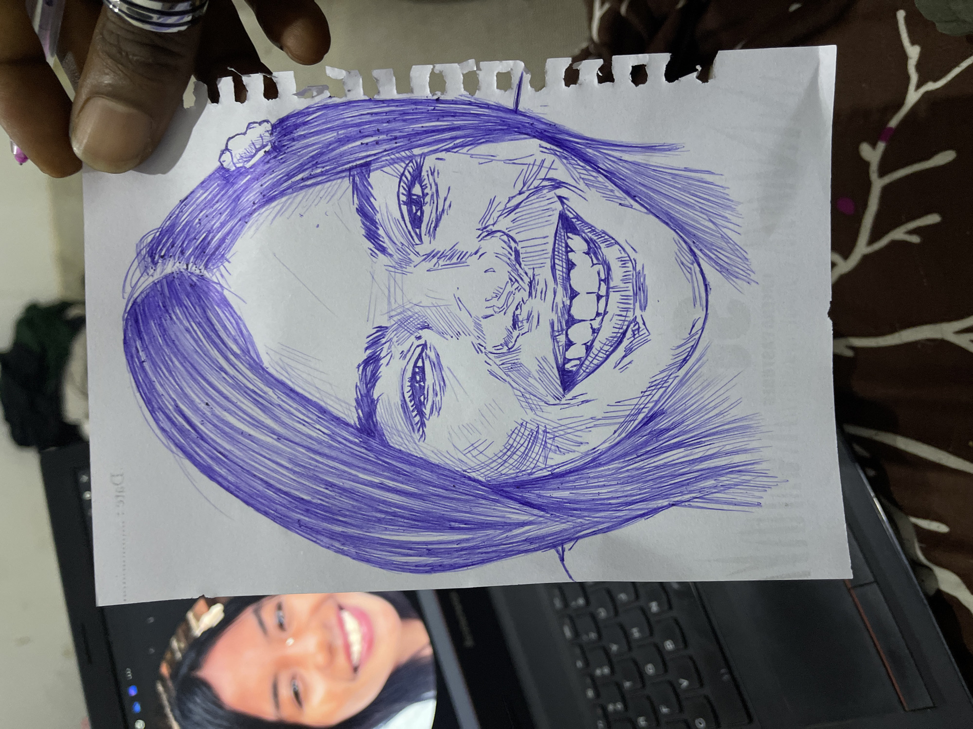 A close-up, eye-level shot shows a ballpoint pen sketch of a woman's face on a ripped piece of white paper. The sketch is done in blue ink and depicts the woman with her mouth open in a wide smile, revealing her teeth. Her eyes are visible, and her hair is drawn with long, flowing lines. A small white flower is sketched in her hair. The paper is held by a person's hand, which is visible in the upper right corner of the image. The person is wearing a ring on their finger. To the left of the sketch, a laptop is partially visible. The screen of the laptop displays a photograph of a woman who appears to be the subject of the sketch. The background of the image is blurred and appears to be a bedspread with a floral pattern. The scene is indoors, suggesting a personal space. The location of Abuja, Nigeria, is not depicted in the image.