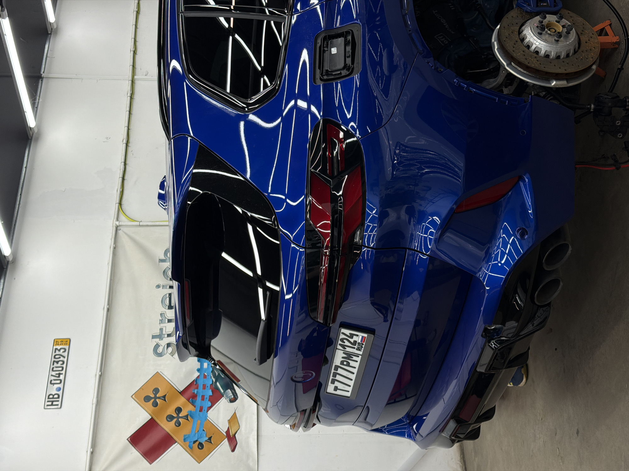 A medium-angle, indoor shot shows the rear of a blue car in a workshop environment. The car has a Russian license plate "T777PM 124 RUS." The rear wheel is removed, exposing the brake disc and caliper. A banner behind the car features an emblem of a cross with orange and red sections, partially covered by blue tape and a plastic bottle. Above the emblem is the partial word "Streich." A Swiss license plate "HB.040393" is affixed to the banner. The location is likely a car repair or detailing shop. The "124" in the Russian license plate suggests the region of Krasnoyarsk, Russia. The lighting is bright, reflecting off the car's polished surface. The background includes equipment and tools typical of an auto repair shop.