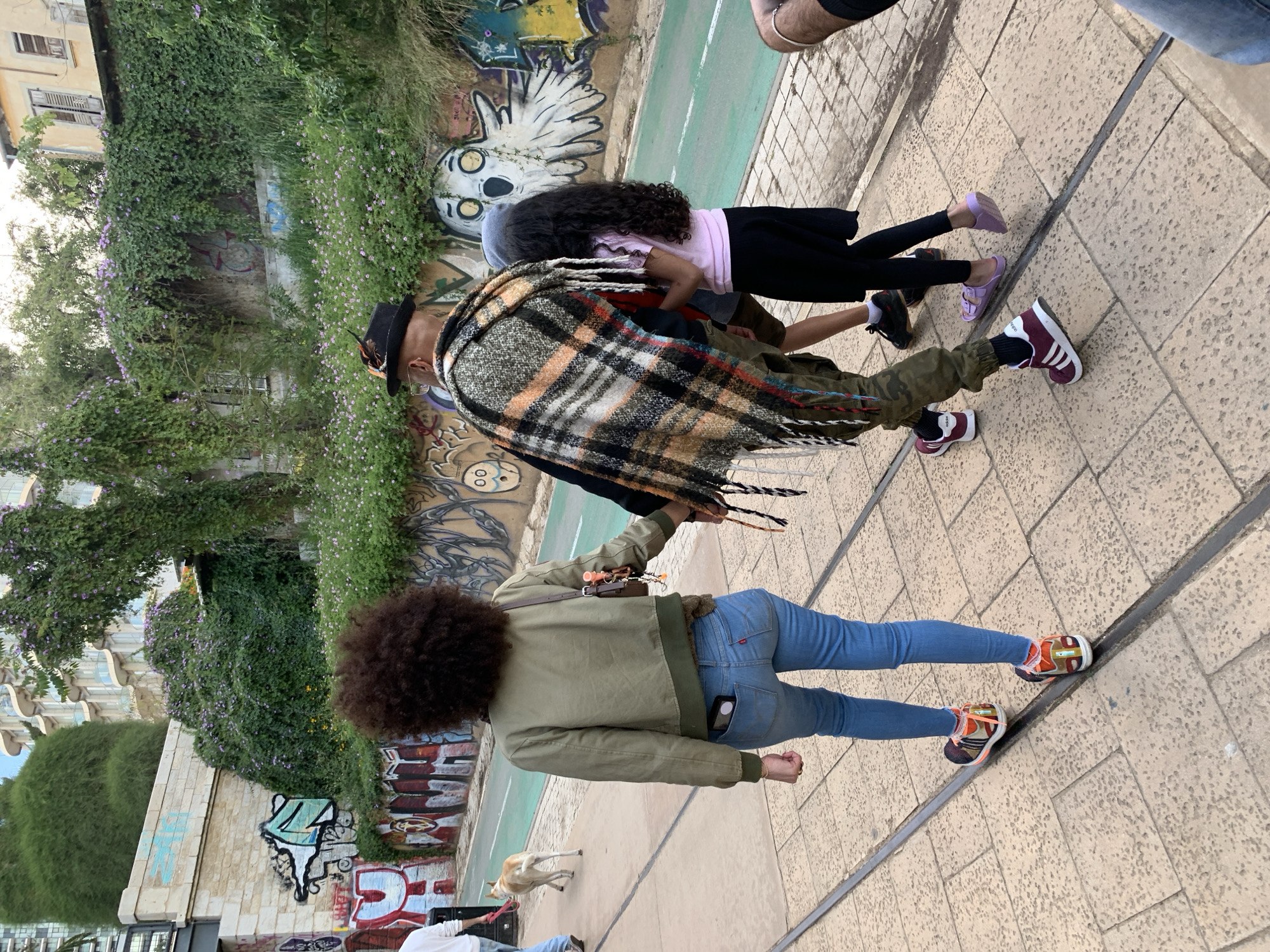 In Tel Aviv, Israel, a group walks on a sidewalk beside a street with graffiti-covered walls. A woman with an afro, wearing a green jacket and blue jeans, is walking to the left of the frame. A man wearing a black hat and a plaid shawl walks next to two children, who are dressed in a pink shirt, black skirt, and sandals. The wall next to them is covered in graffiti and green foliage. A small white dog walks along the street.