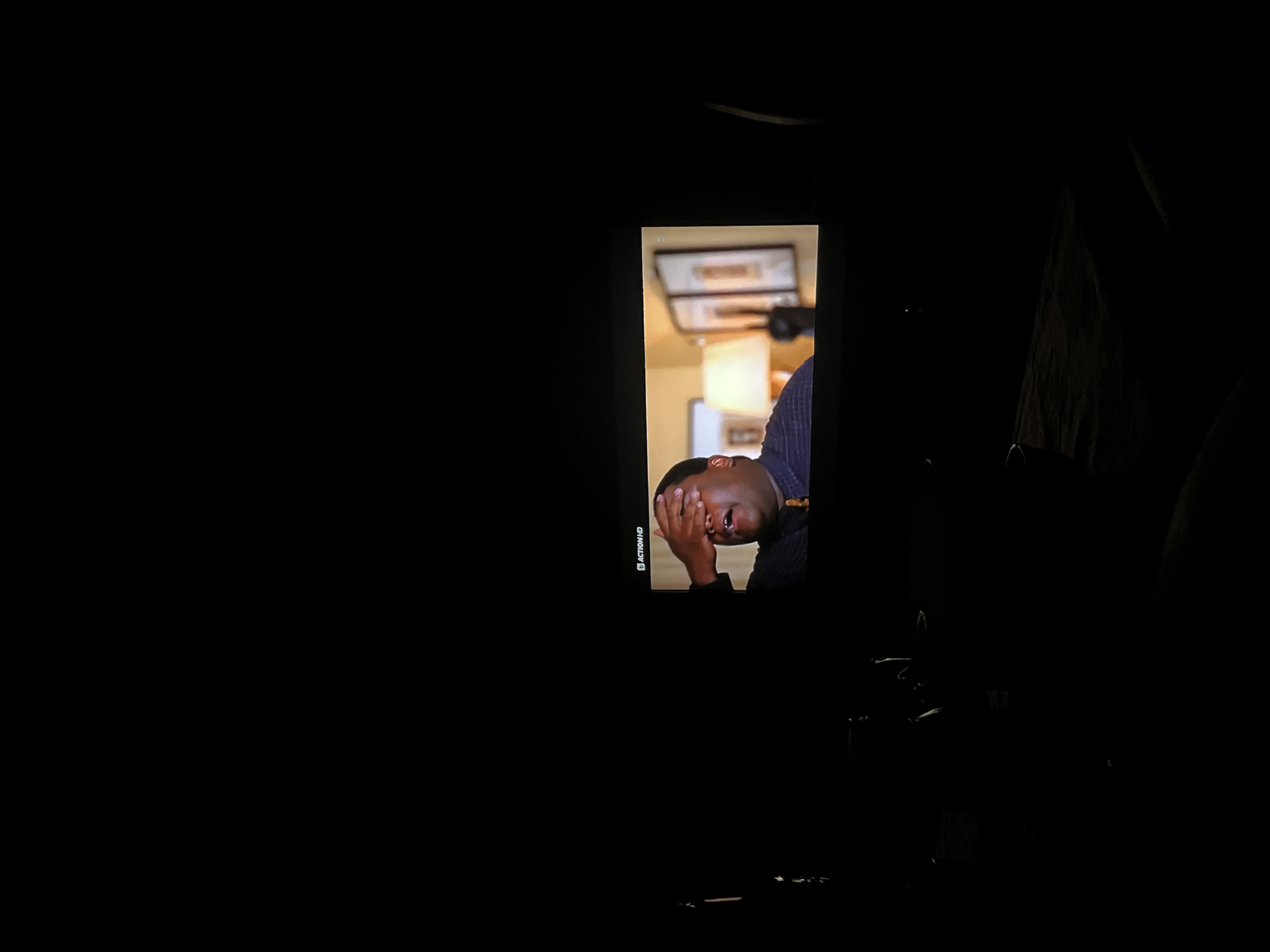 The image shows a dark room with a television screen displaying a scene. On the screen, a Black man is covering his face with his hand, appearing distressed or overwhelmed. The television displays a logo in the top left corner that reads "ACTION HD". In the background of the television screen, a lamp with a yellowish shade is visible, along with two framed pictures on the wall. The rest of the room is mostly obscured by darkness, with some faint shapes suggesting furniture or objects. There is no indication of the location being Plovdiv, Bulgaria; this information is not present in the image.