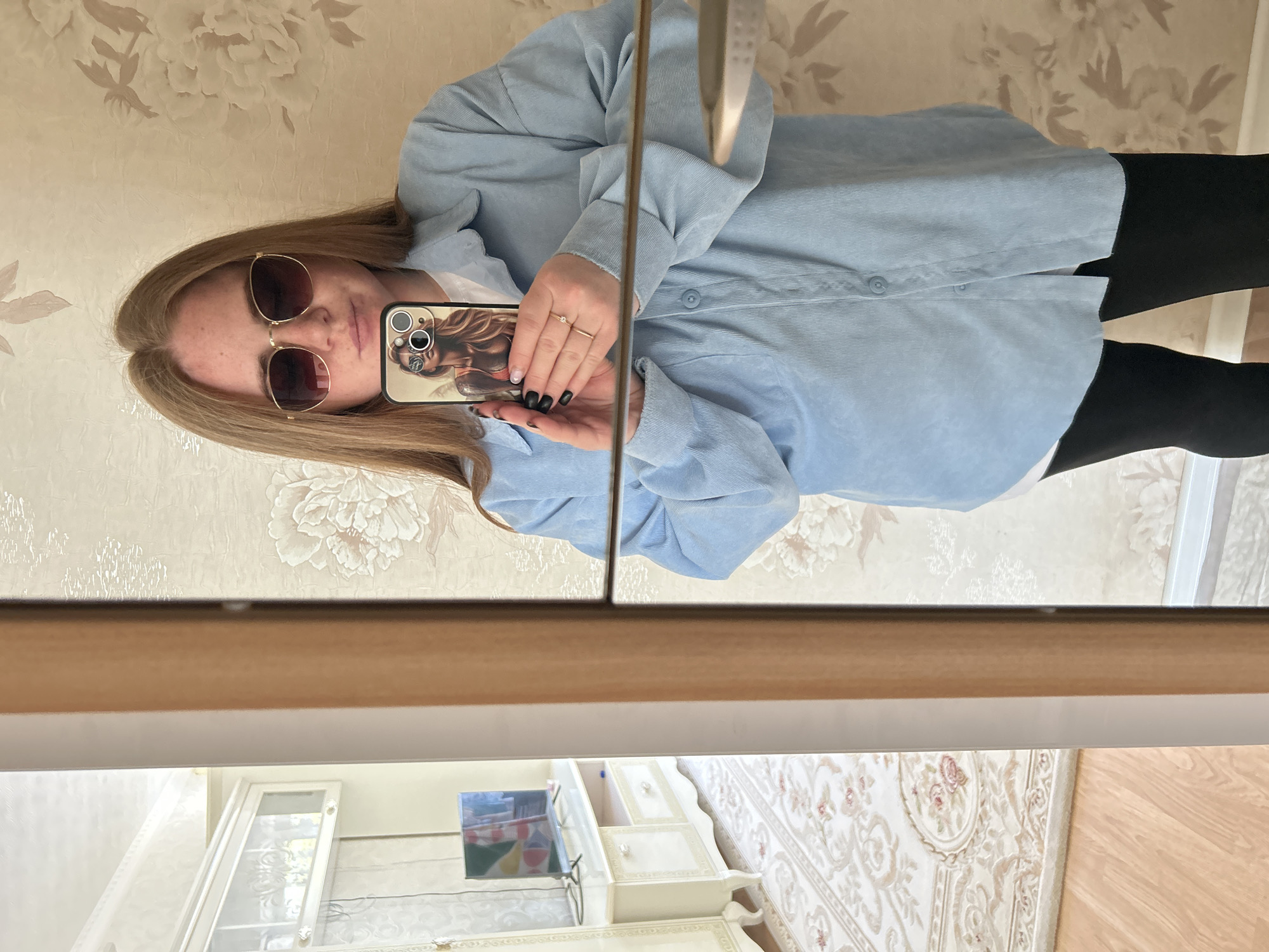 A young woman is taking a selfie in front of a mirror. She is wearing sunglasses, a light blue corduroy button-down shirt, and black pants. She is holding her phone, which has a decorative case with an image of a woman on it. Her fingernails are painted black. The reflection in the mirror shows a room with floral wallpaper, a white cabinet with glass doors, and a television. The room also contains a dresser with decorative handles and a patterned rug. The location is likely in an interior space in Chaikovsky, Russia.