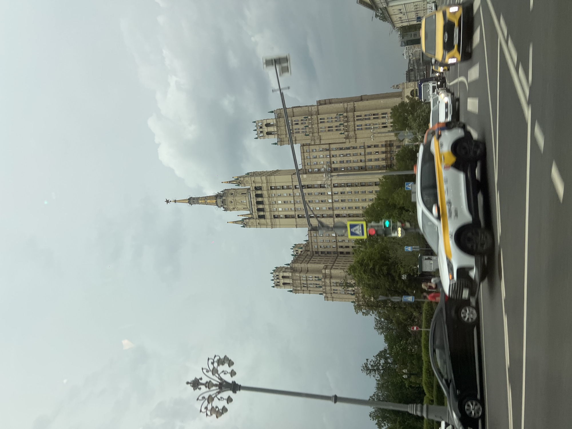 The image shows a view of the Kotelnicheskaya Embankment Building in Moscow, Russia, under a cloudy sky. The building, a Stalinist skyscraper, is centrally positioned with its distinctive spire reaching upwards. A busy city street is visible in the foreground, with several cars in motion. A black sedan and a white and yellow taxi are prominently featured on the road. A vintage-style lamppost stands on the left. Pedestrian crossing signs and a traffic light signal are seen, implying street activity. The blurred motion of the vehicles suggests they are moving.