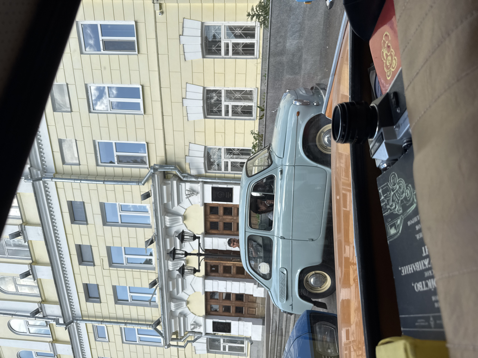The image, taken from inside a vehicle, shows an older blue ZAZ 965 car parked in front of a yellow building in Kazan, Russia. The building is multi-storied and features several windows and a decorative facade with columns and archways above the main entrance. A person is visible peering out of the car's window, while another person stands near the entrance of the building. An old film camera is positioned within the viewing frame of the primary photo. The camera is next to a book that is in a language other than English.
