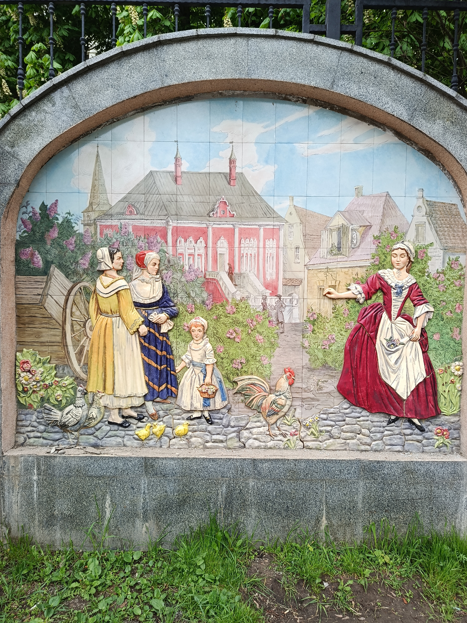 The image depicts a ceramic tile mural set within a stone archway, likely part of a larger structure in Moscow, Russia. The mural depicts a scene from a historical setting, showing figures in what appear to be 18th-century clothing.
On the left, two women in long dresses, one yellow and one blue, stand near a wooden cart. A young girl in a white dress with a blue sash holds a basket. In front of them are several small yellow chicks and a white hen.
To the right, a woman in a red dress with a white apron and trim is standing. Near her is a rooster. In the background is a detailed depiction of a village or town with various buildings, including a red structure with multiple stories and a tall church steeple.
The mural is set against a sky-blue and cloudy background. The archway is made of gray stone. Below the mural, green grass grows from the ground. A metal fence is visible in the background above the archway. The tiles give the impression of depth and relief.