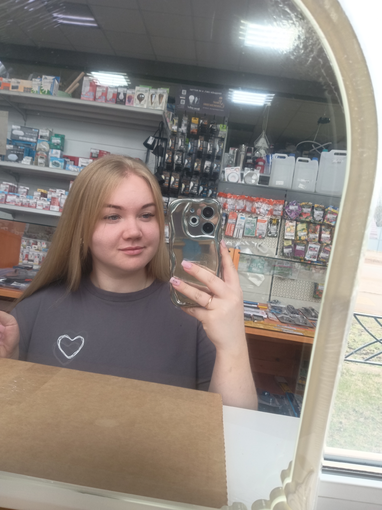 A woman with blonde hair takes a selfie in a mirror. She is wearing a grey t-shirt with a small white heart design. She holds a silver-colored smartphone in her right hand. The reflection shows shelves stocked with various items, including household supplies, light bulbs, and gardening products. The setting is likely a small retail store. The location, Фоки, Russia, is specified, but without additional visual context, this detail doesn't add much to the description.