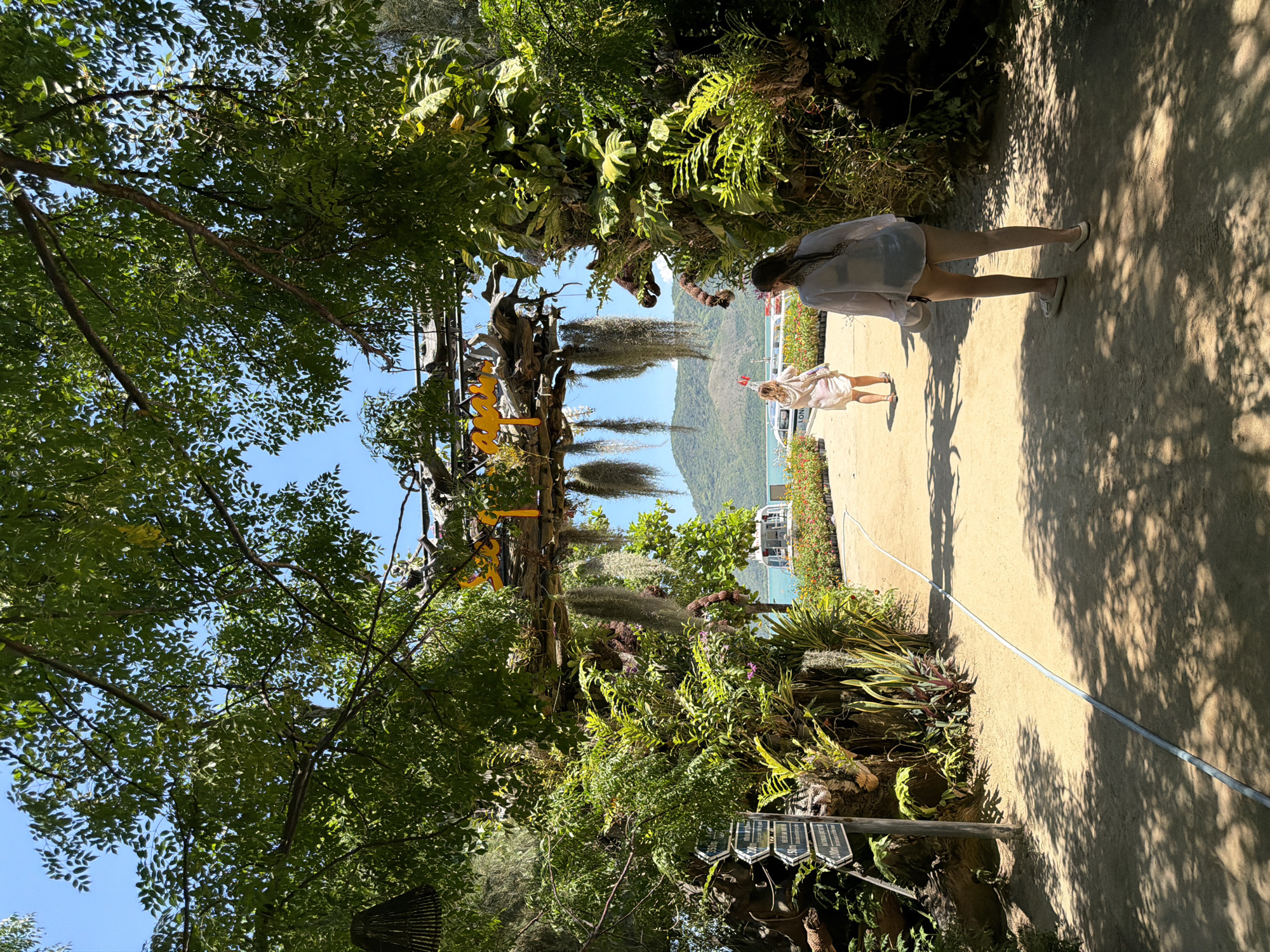 The image shows an outdoor scene with lush greenery and a pathway leading towards a body of water. Two women are walking on the pathway. The woman in the foreground is facing away from the camera, wearing a white shirt and dark bottoms. The woman in the background is also facing away and is wearing a light-colored dress. There are trees with hanging vines and foliage framing the scene. In the distance, there are boats on the water and a mountainous landscape under a clear blue sky. The overall impression is of a tropical or coastal resort area.