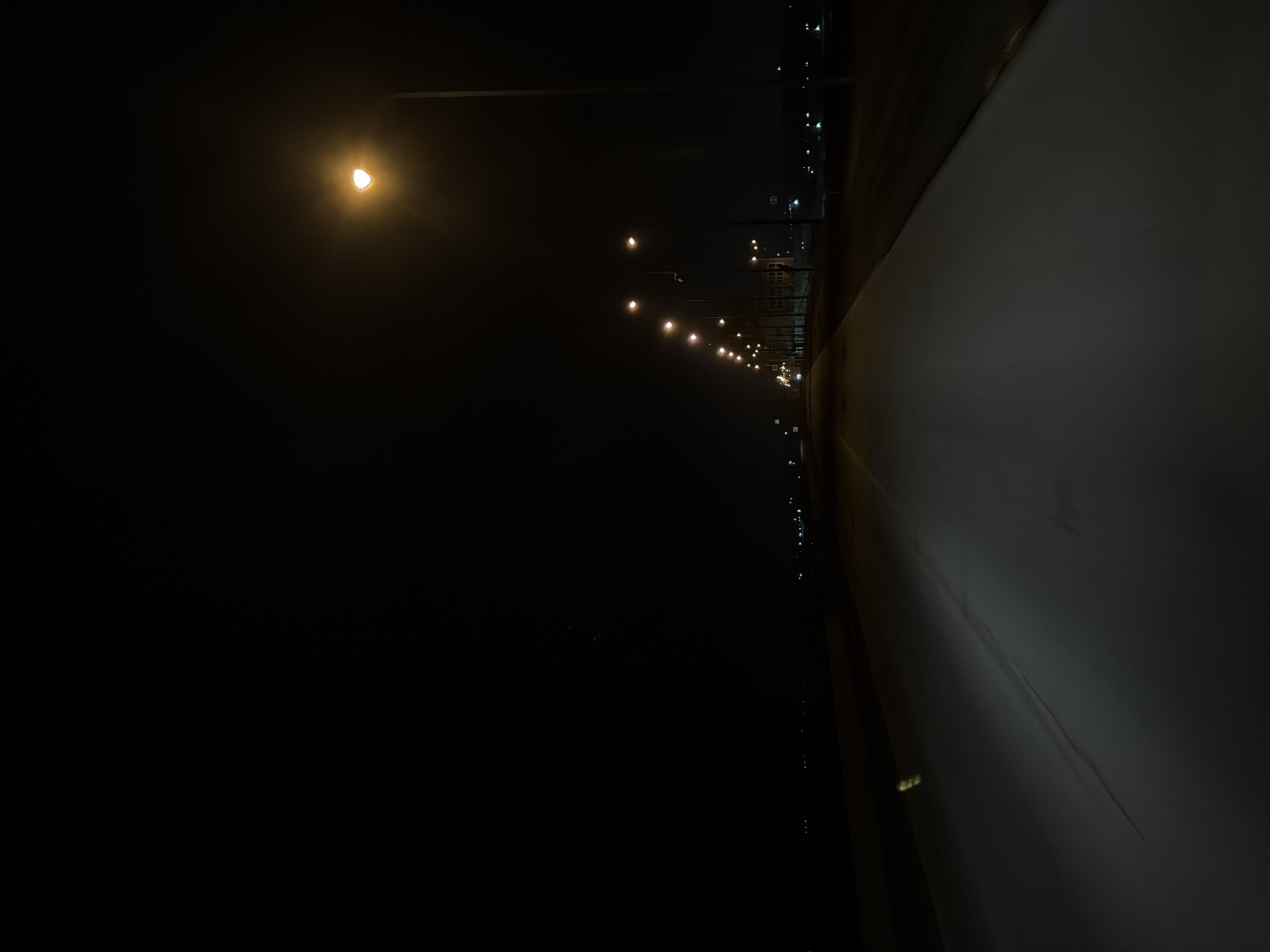 The image depicts a nighttime scene on a road or sidewalk, likely in an urban area. A row of streetlights illuminates the right side of the image, casting a glow on the road surface. The left side of the image is shrouded in darkness. Other buildings are suggested in the distance, faintly lit. The image is taken from a low angle, and is rotated counterclockwise 90 degrees. The location of this image is in the city of Chaikovsky, Russia.
