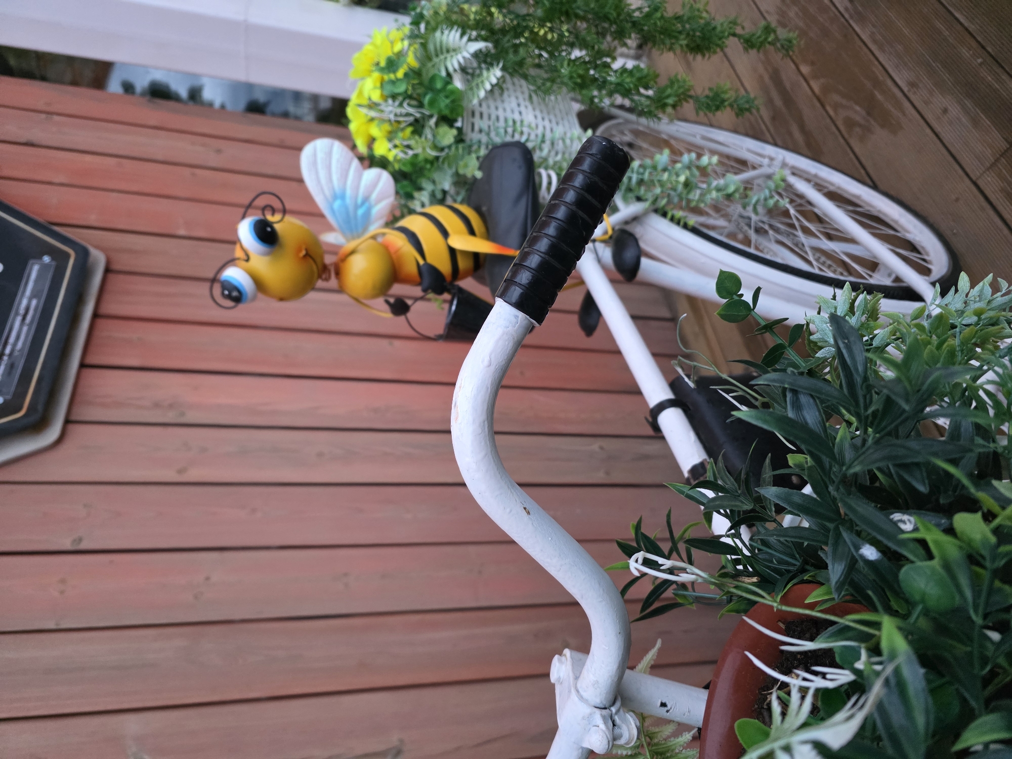 A decorative bicycle is leaning against a wooden wall. A metal bee adorns the wall. The bicycle has artificial plants and flowers in a basket attached to it. The location is Chaikovsky, Russia.