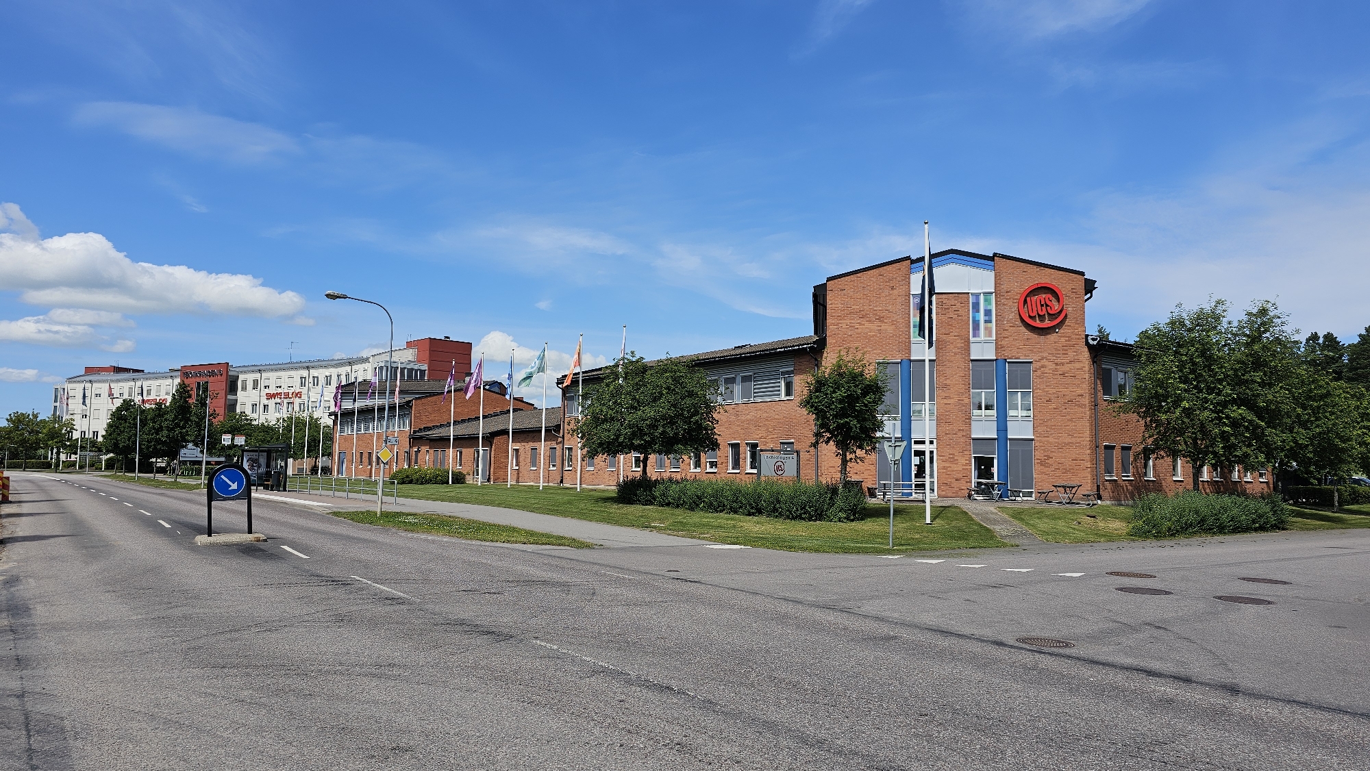 The image shows a wide, paved road curving to the right in the foreground. On the right side of the road is a brick building with a modern blue and gray section on the corner. A large red circular logo with the letters "UCS" in white is visible on the upper part of the brick building. There are several flags on poles in front of the building. To the left of the road, there is a sidewalk, a bus stop shelter, and a row of multi-story buildings in the background. The sky is blue with scattered white clouds. The scene appears to be an industrial or commercial area in Linköping, Sweden.