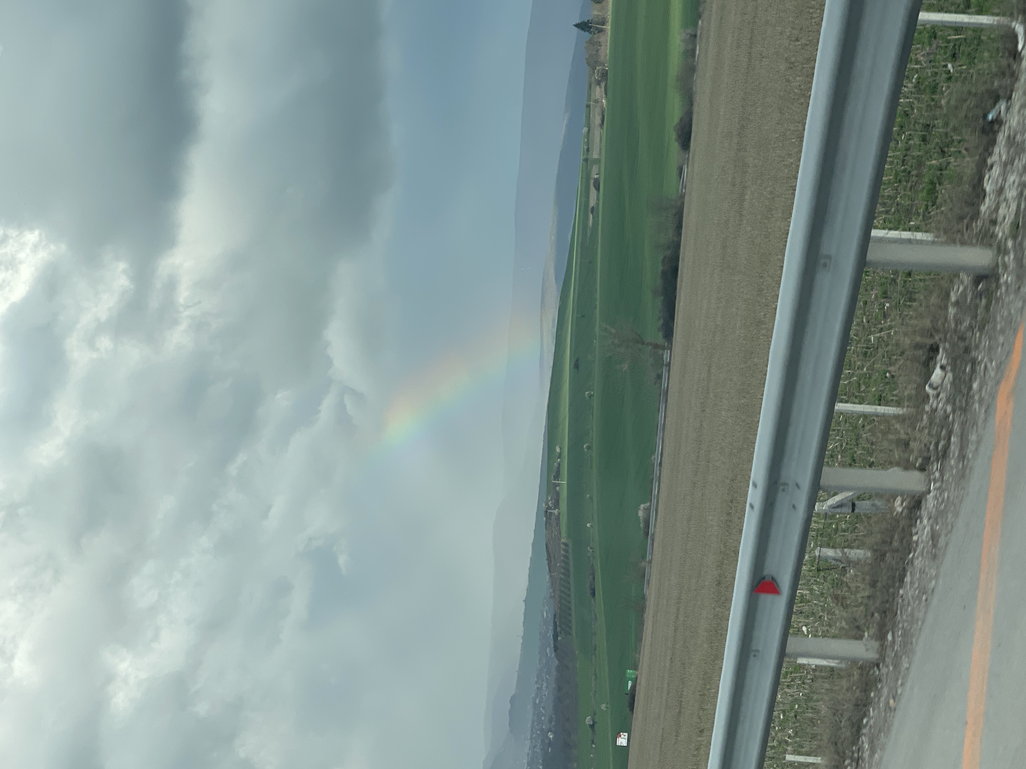 A landscape scene shows a rainbow in a cloudy sky over a rural area, possibly near Buhovtsi, Bulgaria, as suggested by the location. A gray metal guardrail, with a red reflector, is on the right side of the image. Behind the guardrail are fields. Patches of green grass are visible along the edge of the road, beyond the guardrail. Further beyond, a mix of green and tan fields extends into the distance towards some mountains. The rainbow is on the left side of the image in the distance. The sky is predominantly covered with gray clouds.