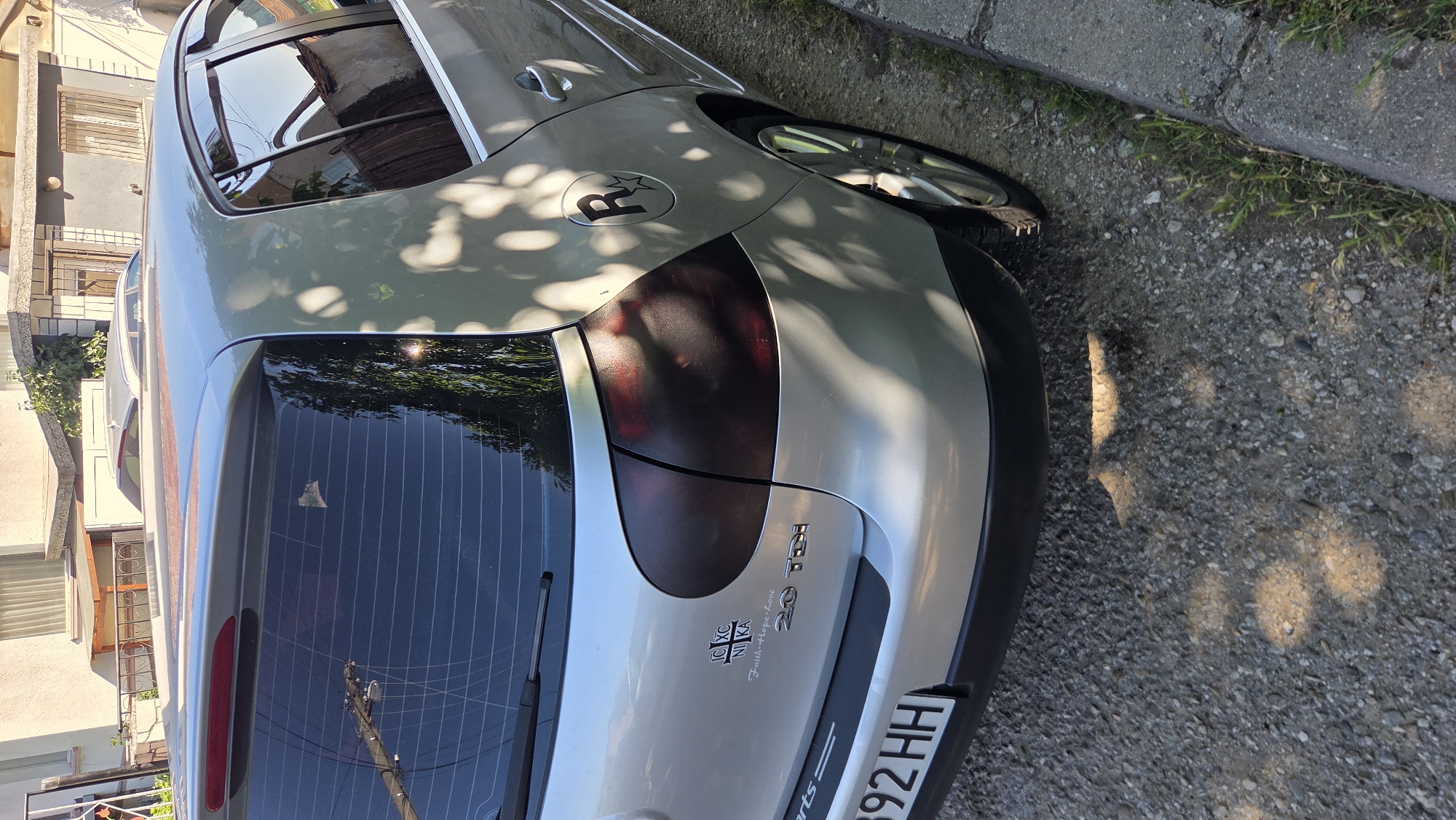 A silver Volkswagen Golf is parked on a gravel surface next to a concrete curb in an urban environment. The car's rear is facing the camera, and the license plate reads "892 HH". A "2.0 TDI" emblem is visible on the trunk. There is a decal on the car with Greek letters and the words "Faith-Hope-Love". A circular sticker with the letter "R" and a star is on the fuel door. Sunlight casts dappled shadows of leaves on the car's surface, suggesting the presence of trees or foliage nearby. The background shows residential buildings with windows and roofs. The overall scene is outdoors and appears to be during daylight hours.