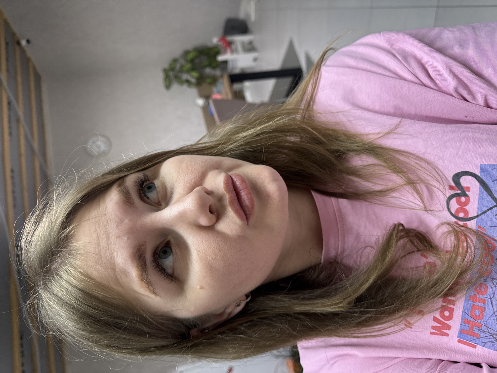 A close-up portrait depicts an adult female indoors in Gornyi (Dvurechenskoe S/P), Russia. The subject has long, light brown hair. Her blue eyes are directed upwards and to the viewer's left. Her lips are slightly pursed. A small, light brown mole is visible on her right cheek (viewer's left). She wears a small hoop earring in her left earlobe and a stud in the upper cartilage. Her attire consists of a pink long-sleeved garment with partial text reading "Wan" and "I Hate" visible on the chest area. The background, blurred, shows a light-colored wall, a suspended ceiling with dark supports, and a white-faced wall clock. A small, dark security camera is mounted near the ceiling. Further back, a green potted plant, a wooden table with a dark frame, and white chairs are visible. The floor appears tiled.