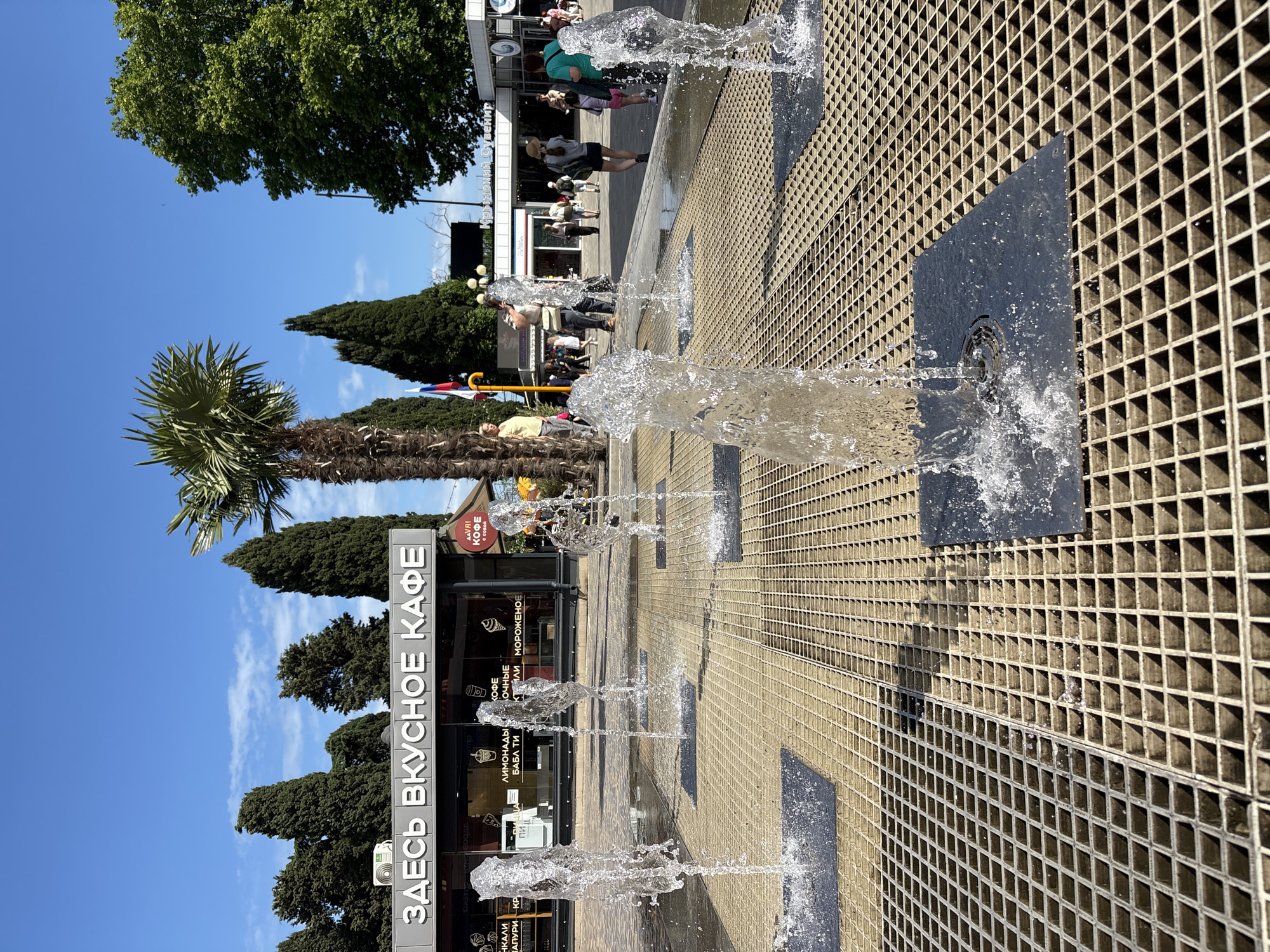 The image shows a street scene with a ground-level fountain in the foreground. The fountain consists of multiple jets of water spraying upwards from square metal plates set within a grid-like metal surface. In the background, there is a cafe with a sign reading "ЗДЕСЬ ВКУСНОЕ КАФЕ" (Here is a tasty cafe). Behind the cafe, there are people walking and trees. A palm tree is visible to the right of the cafe. The sky is blue with some clouds. The scene appears to be a public area, possibly a promenade or square. The location is identified as Alushta, Ukraine, although this information cannot be independently verified from the image alone.