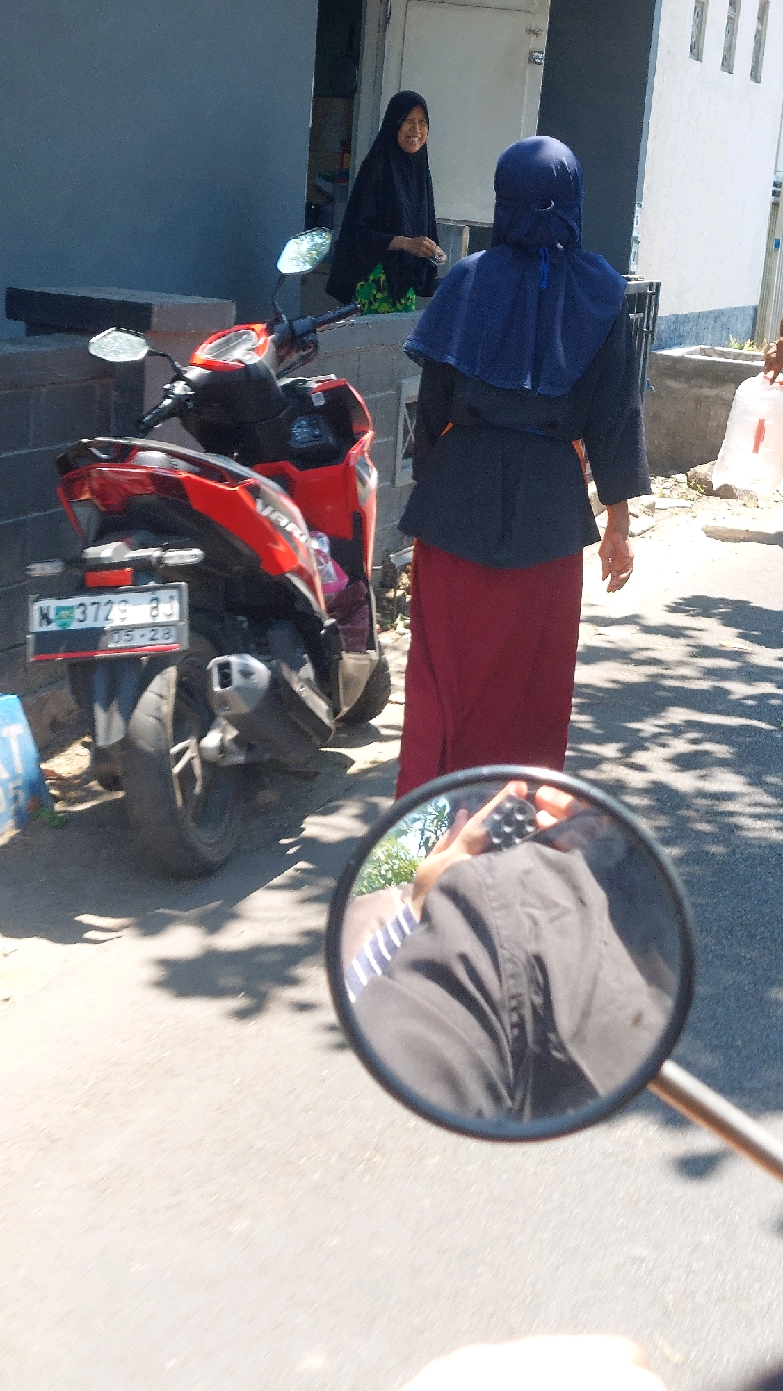 In Siwalan Panji, Indonesia, a red and black scooter is parked on a paved surface. A woman wearing a black hijab and a green patterned skirt stands in the background near a doorway, smiling. In the foreground, a second woman, also wearing a hijab, is seen from the back, dressed in a dark blue veil over a dark jacket and a maroon skirt. Her right hand is visible, and she appears to be holding onto the handlebars of a motorcycle, as evidenced by the reflection in the motorcycle's rearview mirror, which shows part of her hand and a black and white striped garment. The environment is outdoors, with sunlight casting shadows on the ground, suggesting it is daytime. A building with a white wall and barred windows is visible to the right.