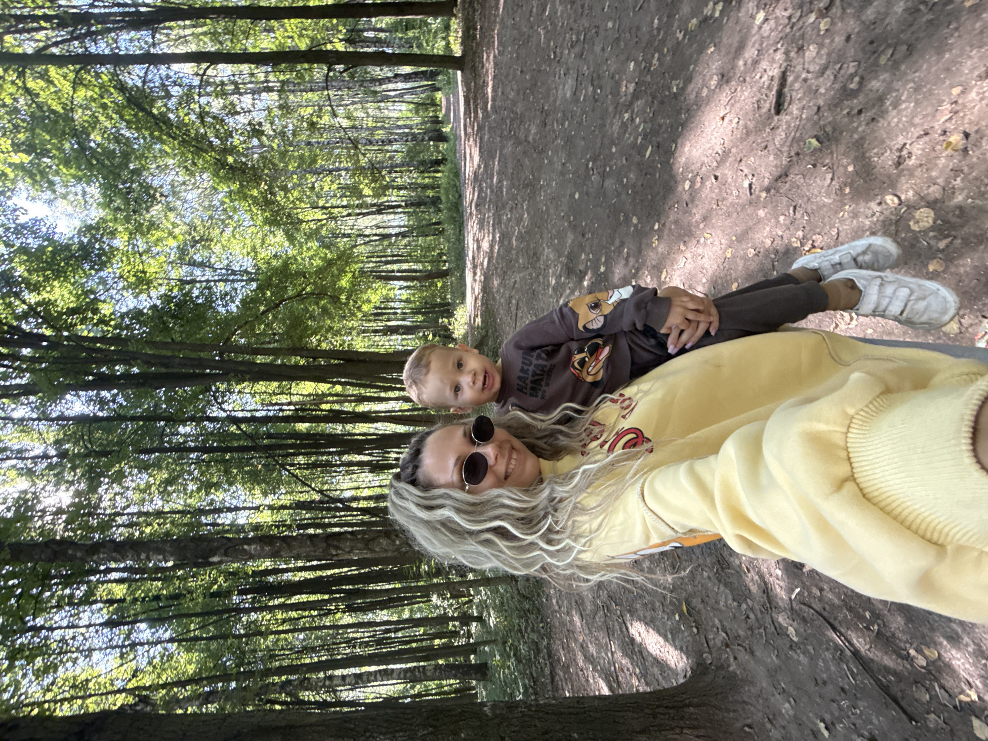 A medium-shot, waist-up selfie shows a woman and a young child posing in a sun-dappled forest. The woman, with curly blonde hair and dark sunglasses, smiles at the camera while holding the child. She wears a yellow sweatshirt with an orange and red graphic design. The child, with light brown hair, wears a dark brown sweatshirt featuring characters from The Lion King and dark pants. Both are wearing white sneakers. The forest floor is covered with dirt and scattered leaves. Tall trees with green foliage create a canopy overhead, allowing sunlight to filter through. The background shows a path leading deeper into the woods. The location is identified as Dumino, Russia.