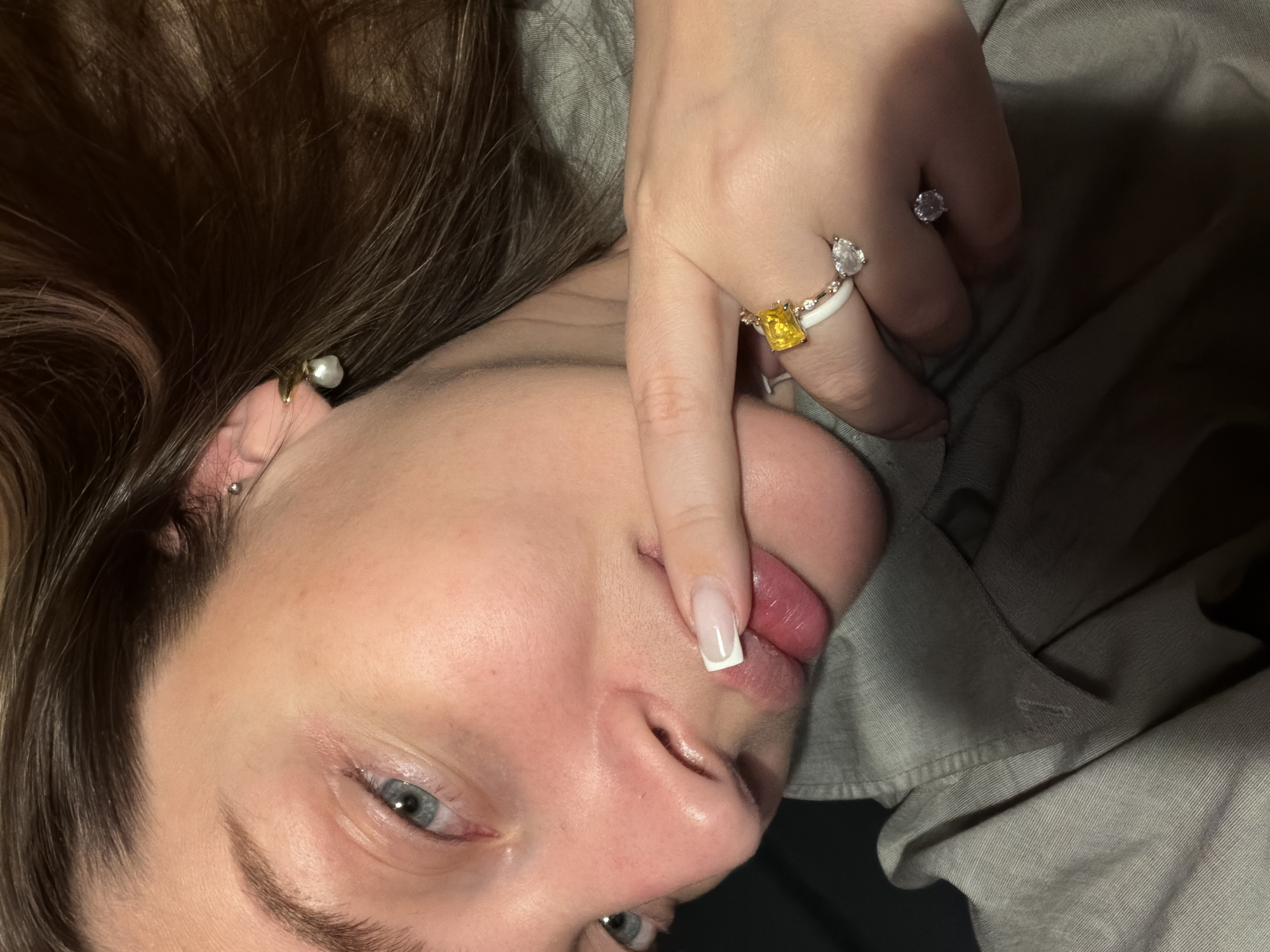 The image shows a fair-skinned individual with brown hair, light blue eyes, and a French manicure. The person is wearing multiple rings on their fingers, one with a yellow stone and another with a clear stone. One finger is touching their lips. A pearl earring and a stud earring are visible on the left ear. The person is wearing a light gray shirt. There is no information given in the image to suggest the location is Moscow, Russia.